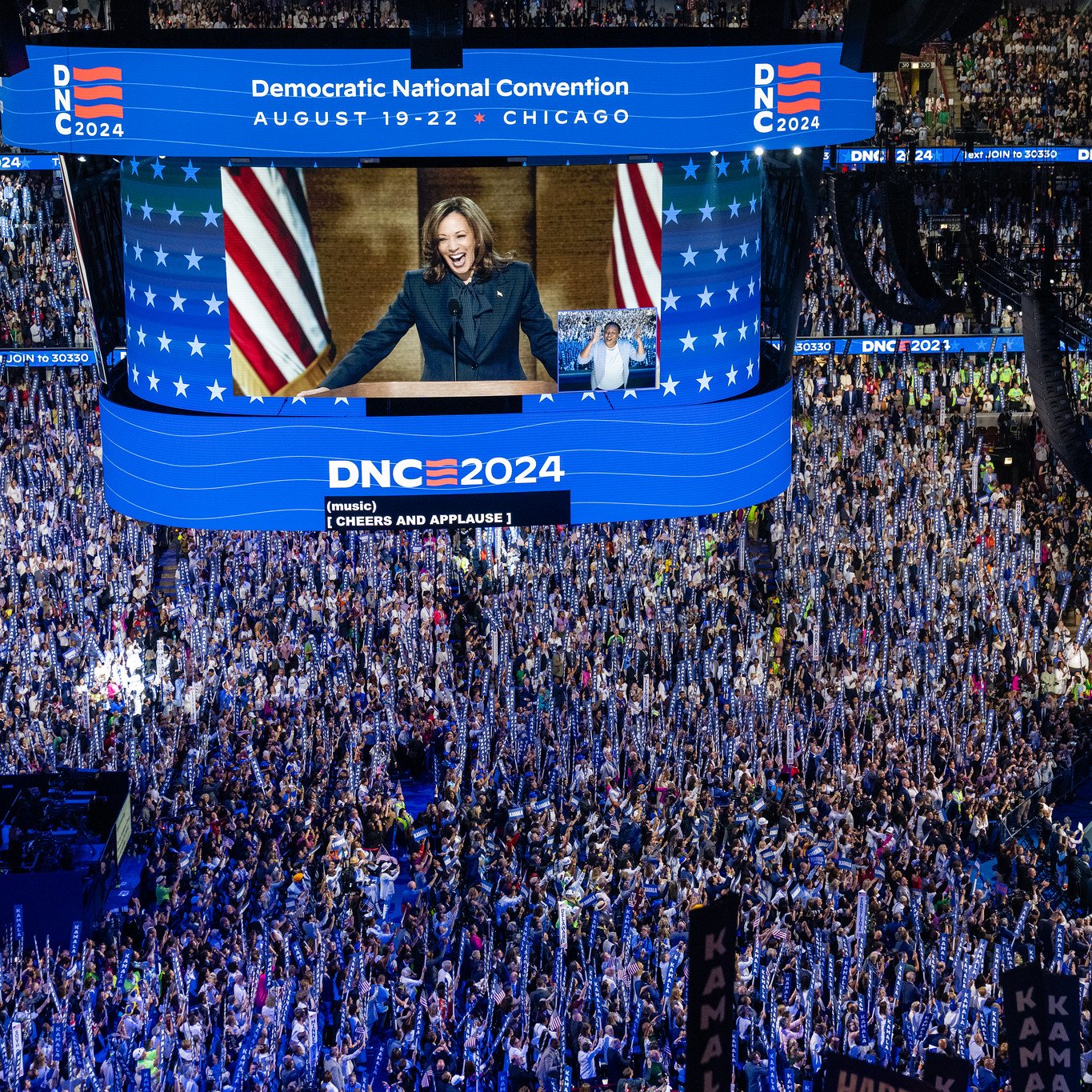 L'immagine mostra una scena di una grande convention politica, probabilmente la Convenzione Nazionale Democratica del 2024 a Chicago. La sala è affollata di persone che applaudono, con molti sostenitori che sventolano bandiere e cartelli. Al centro, un grande schermo mostra un discorso di una figura politica (probabilmente un leader del partito). Gli ambienti sono decorati con colori e simboli del partito, come stelle e strisce. L'atmosfera sembra essere di entusiasmo e impegno politico.