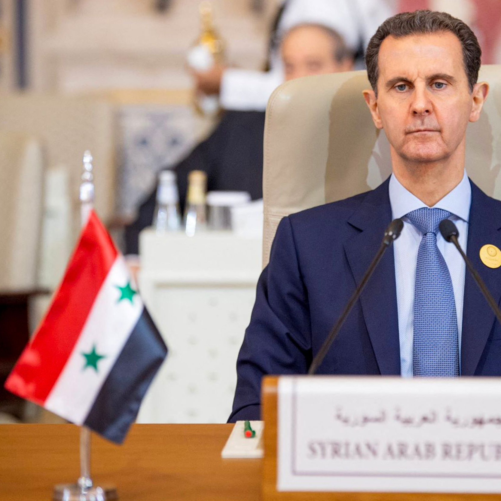 در این تصویر، فردی در یک نشست رسمی نشسته است. او کت و شلوار تیره‌ای به تن دارد و به دوربین نگاه می‌کند. در جلوی او، یک تابلو با نوشته «جمهوری عربی سوریه» و پرچم سوریه قرار دارد. در پس‌زمینه، تعدادی از افراد دیگر نیز در حال حضور در جلسه هستند. فضای تصویر به نظر جدی و رسمی می‌آید.