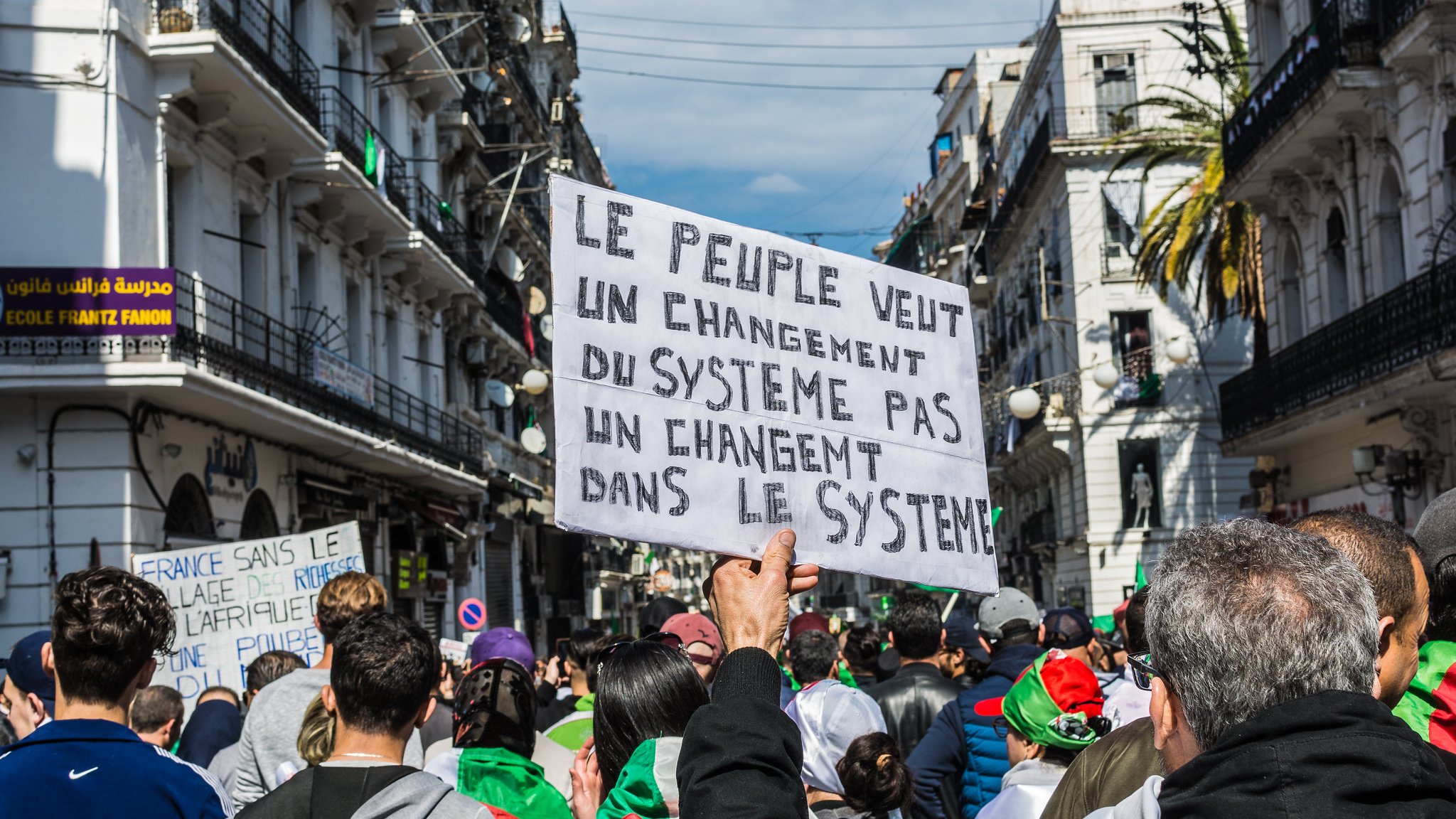 L'image montre une manifestation dans une rue animée. Au premier plan, une personne tient une pancarte qui exprime un message clair : "Le peuple veut un changement du système pas un changement dans le système". On aperçoit des manifestants qui portent des drapeaux et semblent mobilisés. Les bâtiments environnants évoquent une ambiance urbaine et dynamique, tandis que le ciel est partiellement nuageux. L'ensemble traduit une forte volonté collective de changement.