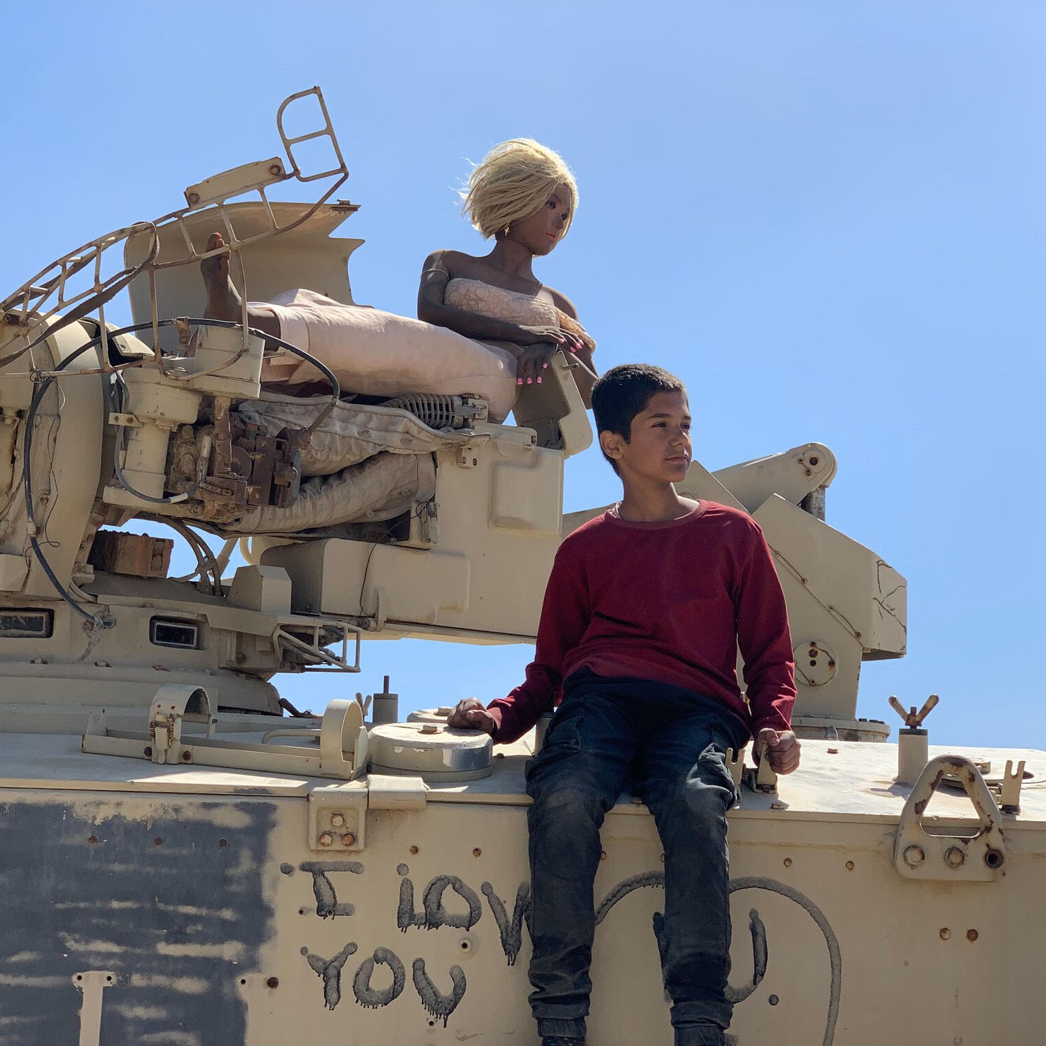 L'immagine mostra un giovane ragazzo seduto su un veicolo militare, apparentemente un carro armato. Accanto a lui c'è una figura che sembra essere una bambola o un manichino con capelli biondi. Sul carro armato è visibile una scritta che dice "I love you". Il cielo è limpido e blu, creando un contrasto con l'ambiente circostante. Il ragazzo indossa un maglione rosso e jeans, mentre la bambola è posizionata in modo da sembrare tranquilla e serena.