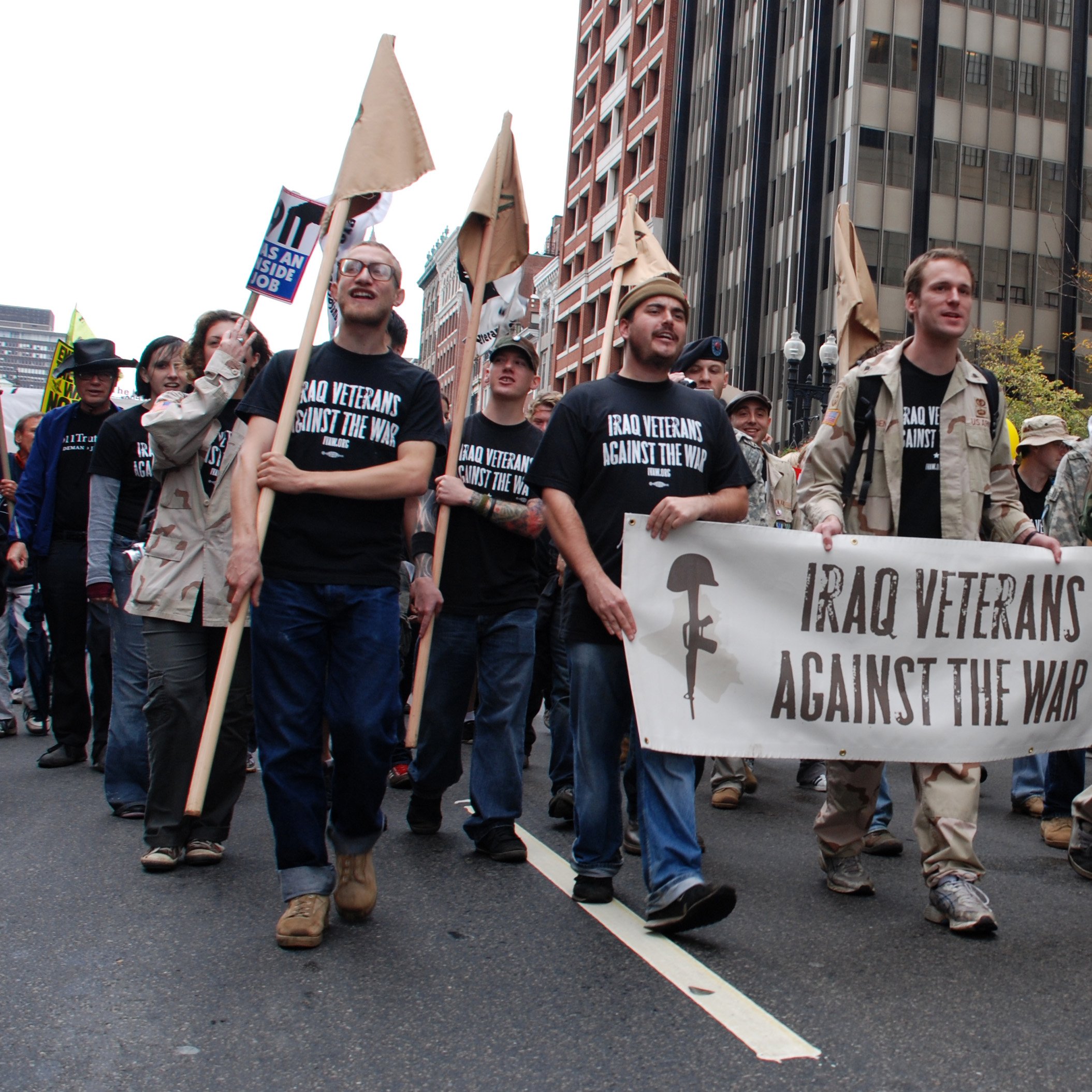 L'image montre un groupe de manifestants marchant dans une rue, tenant des pancartes. Au premier plan, plusieurs personnes portent des t-shirts noirs avec des slogans, et l'un d'eux tient une grande banderole qui dit "Iraq Veterans Against The War". Il semble s'agir d'une manifestation contre la guerre en Irak, avec des participants d'âges et d'apparences variés. L'ambiance semble sérieuse et engagée. Des bâtiments urbains et des arbres aux feuilles jaunes sont visibles en arrière-plan, suggérant que la scène se déroule en automne.