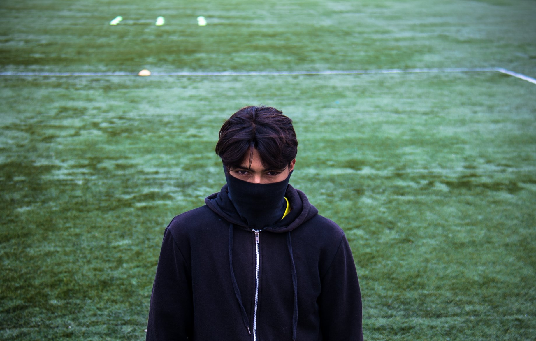 L'image montre un jeune homme se tenant sur un terrain de sport couvert d'herbe. Il porte un sweat à capuche noir et un masque noir qui couvre une partie de son visage. Son regard est intense et fixé vers l'objectif, créant une atmosphère mystérieuse. Le terrain derrière lui est bien entretenu, avec des marques blanches au sol.