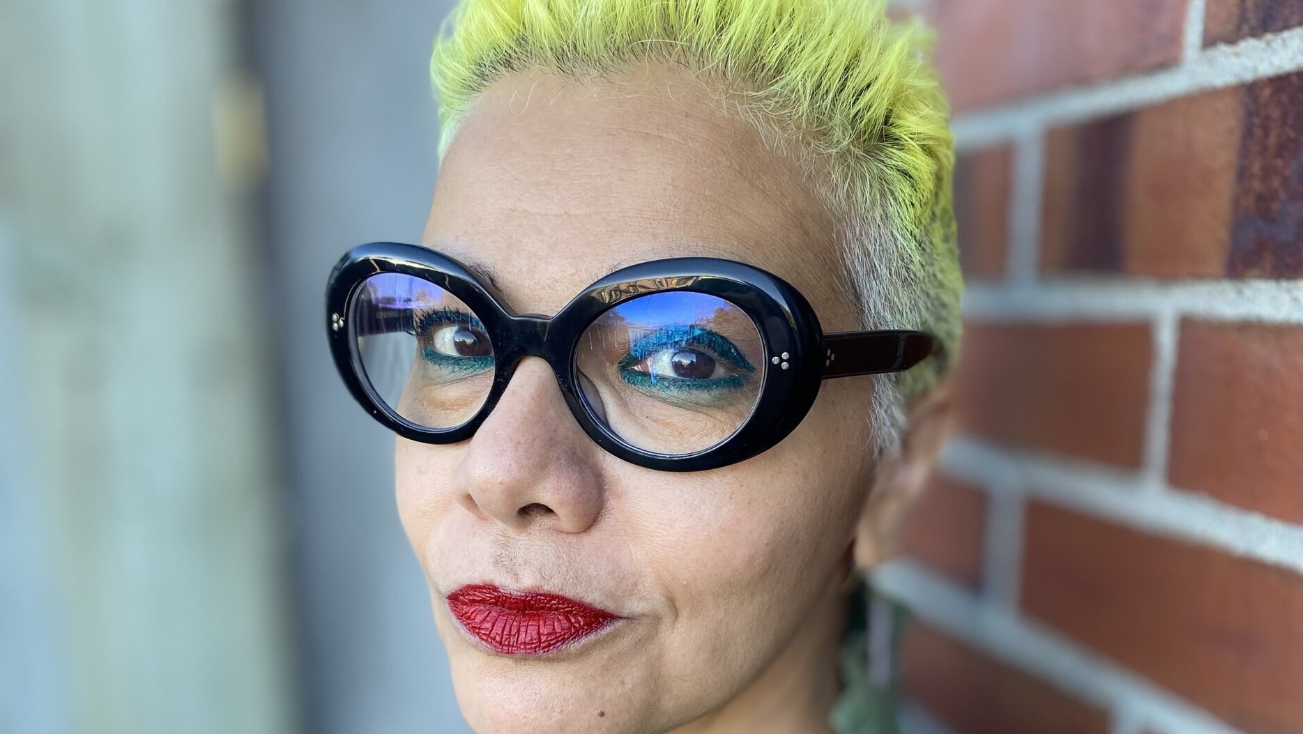 L'immagine mostra una persona con capelli corti e di colore giallo brillante. Indossa occhiali grandi e neri, che aggiungono un tocco audace al suo look. Il trucco è accentuato con labbra rosse, mentre lo sfondo presenta una parete di mattoni, creando un contrasto interessante con i colori vivaci della persona. La sua espressione è sicura e affermativa.