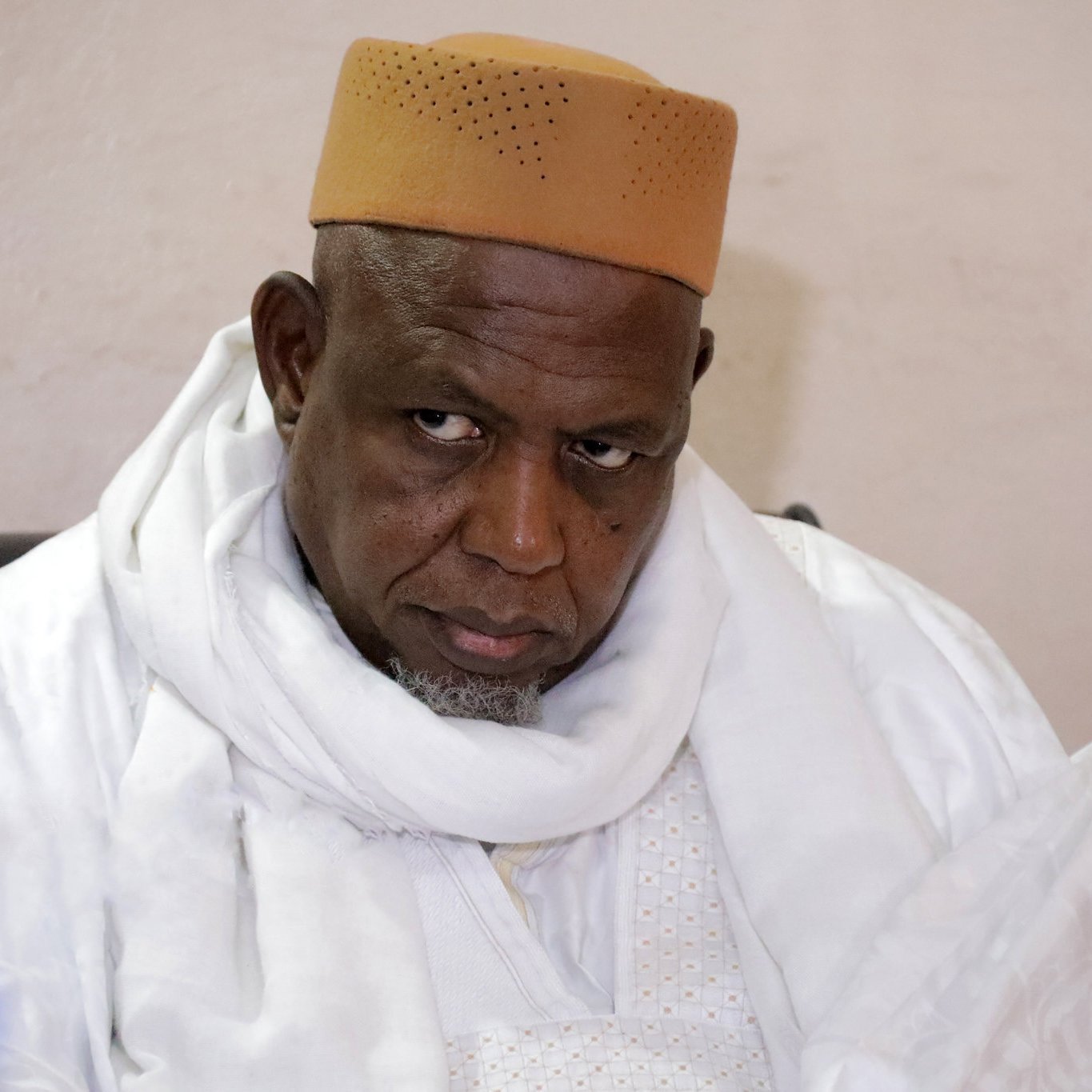 L'image montre un homme, probablement assis, portant une tenue traditionnelle, incluant un chapeau de couleur beige et un long habit blanc. Son expression semble sérieuse et réfléchie, tandis qu'il regarde dans une direction avec une certaine intensité. En arrière-plan, on peut voir des bouteilles d'eau sur une table. L'environnement semble simple, suggérant un cadre formel ou communautaire.