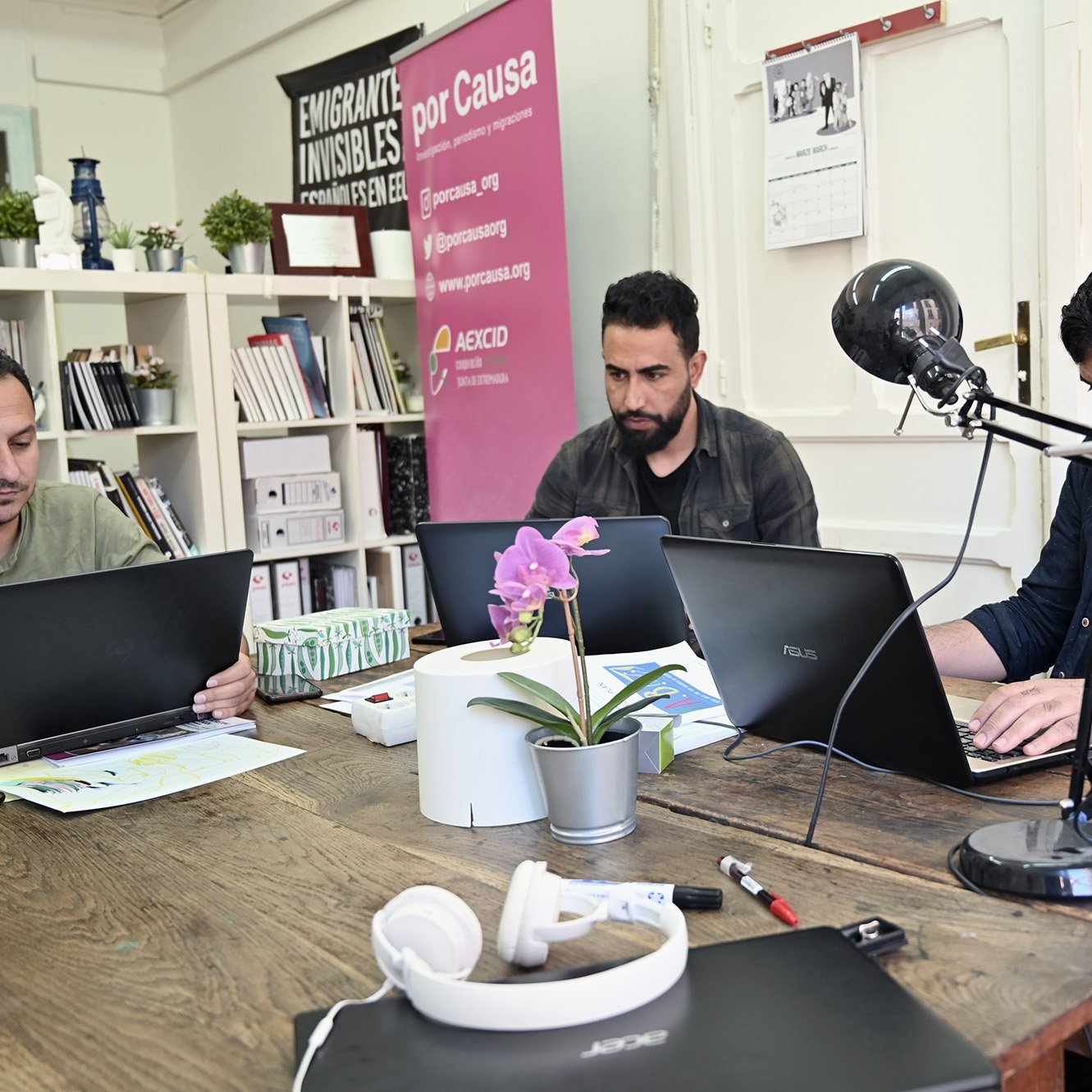 En la imagen se pueden ver a tres hombres sentados en una mesa de trabajo. Cada uno tiene un portátil frente a él y parece que están concentrados en sus tareas. La mesa está decorada con una planta en una maceta y hay auriculares sobre la superficie. En el fondo, hay una pared con algunos carteles, incluyendo uno que dice "Emigrantes Invisibles". También hay estantes con libros y otros materiales. La atmósfera sugiere un ambiente de trabajo colaborativo y dinámico.