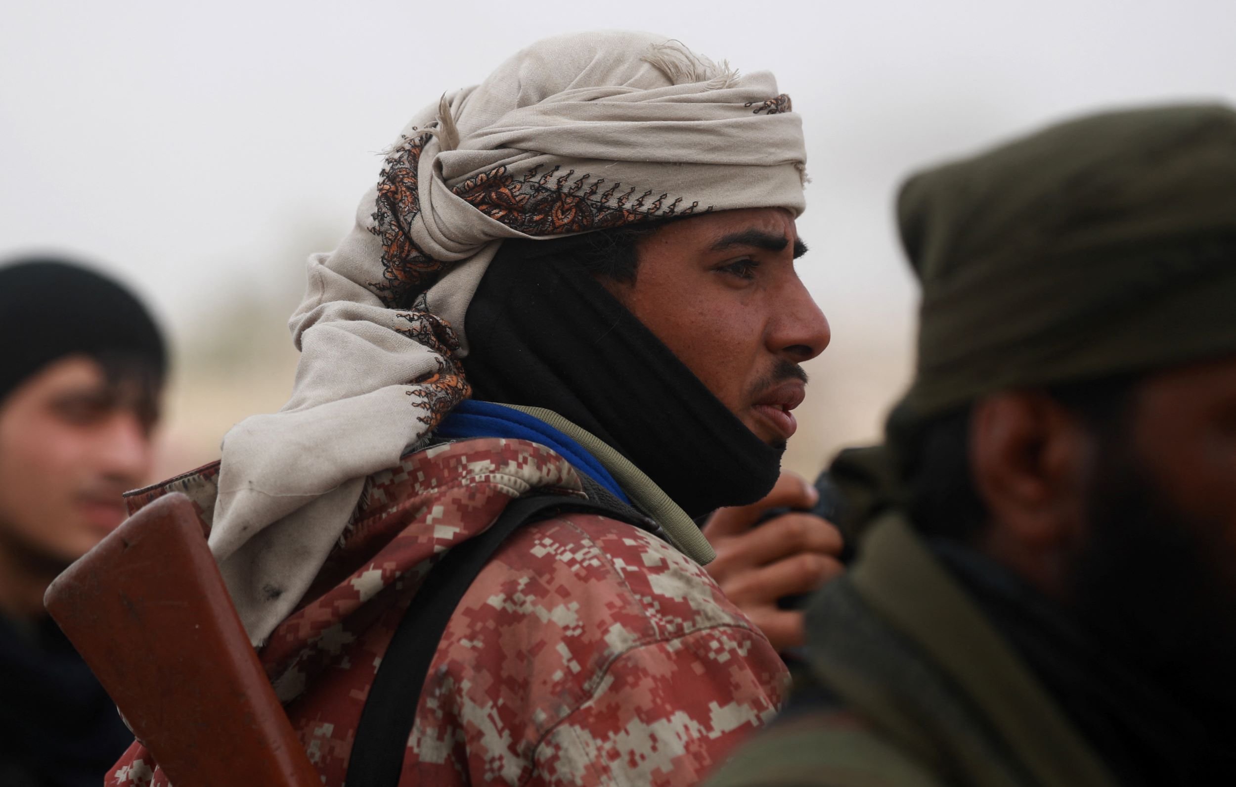 در این تصویر، یک فرد در حالتی نزدیک به دوربین قرار دارد. او کلاهی بر سر دارد و چهره‌اش جزئیاتی را نشان می‌دهد که به او ظاهری خاص می‌دهد. لباسش نیز شامل یک روپوش است که الگوهای خاصی دارد. در پس‌زمینه، افرادی دیگر نیز دیده می‌شوند. جو تصویر احتمالاً جدی و نظامی است.