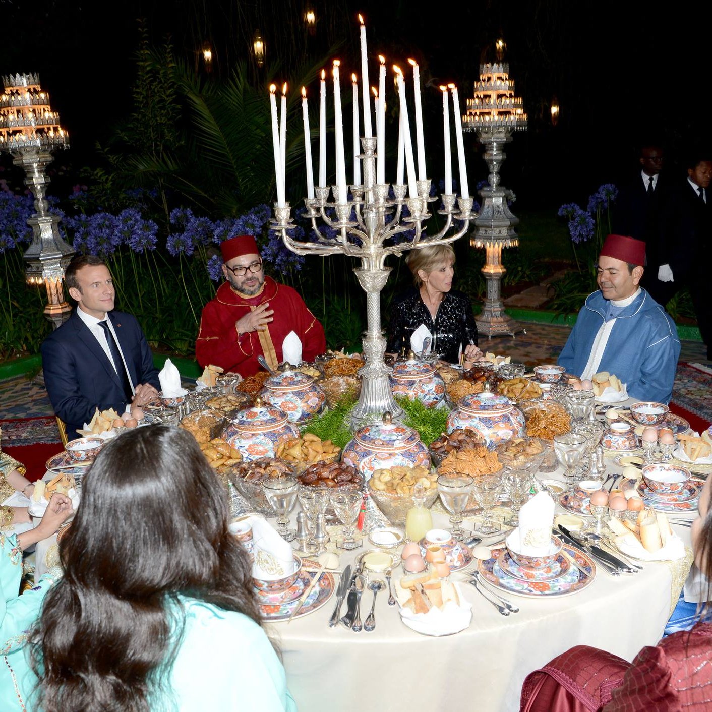 تظهر الصورة مجموعة من الأشخاص حول مائدة رائعة مُعدّة لوجبة طعام فاخرة. المائدة مزينة بأطباق متعددة تحتوي على أطعمة متنوعة، وتحيط بها شمعدانات كبيرة وأجواء أنيقة. يرتدي بعض الضيوف ملابس تقليدية مغربية، بينما يرتدي آخرون ملابس رسمية. تبدو الصورة وكأنها تُعبر عن احتفال خاص أو مناسبة اجتماعية، مع خلفية من النباتات والزهور التي تضفي جوًا جميلًا.