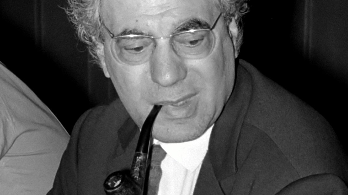 L'image montre un homme assis avec une pipe dans la bouche. Il porte une veste, une chemise blanche et une cravate. Ses cheveux sont grisonnants et légèrement bouclés. Il a des lunettes et semble concentré sur une conversation ou une discussion. En arrière-plan, on peut apercevoir d'autres personnes, mais elles ne sont pas clairement visibles. L'éclairage est modéré, ce qui donne une ambiance sérieuse à la scène.