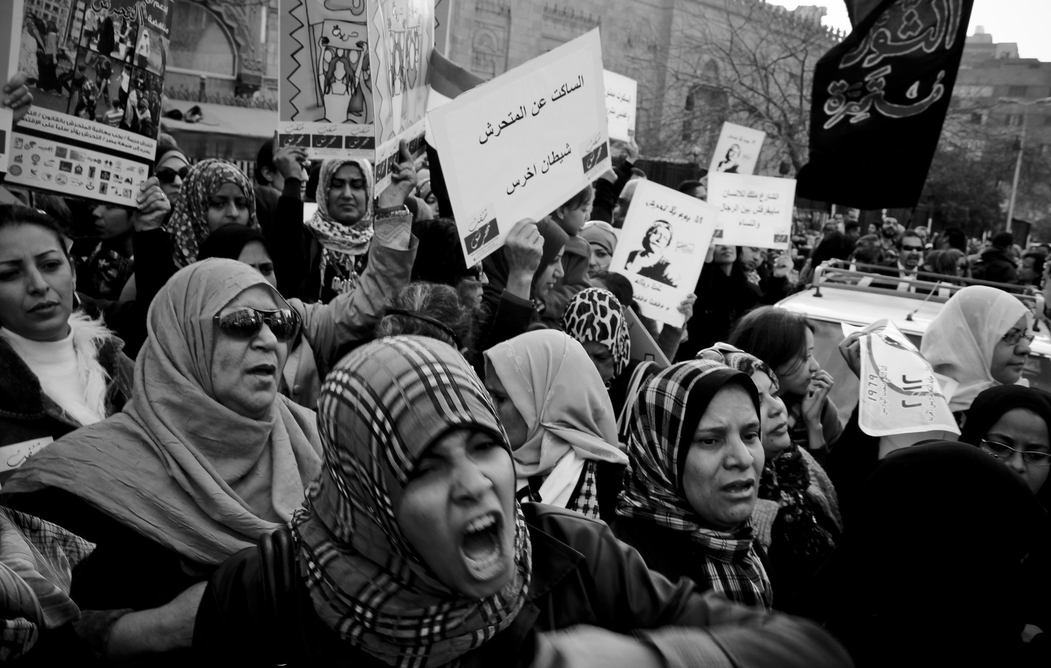 L'immagine mostra una folla di donne che partecipano a una manifestazione. Sono vestite con abiti tradizionali e portano foulard. Alcune di loro sembrano molto appassionate, con espressioni di intensità sul volto. Teniamo in mano cartelli con messaggi scritti, mentre sullo sfondo si possono intravedere altri manifestanti e bandiere. L'atmosfera è di grande mobilitazione e determinazione. La foto è in bianco e nero, il che accentua le emozioni e il senso di protesta.
