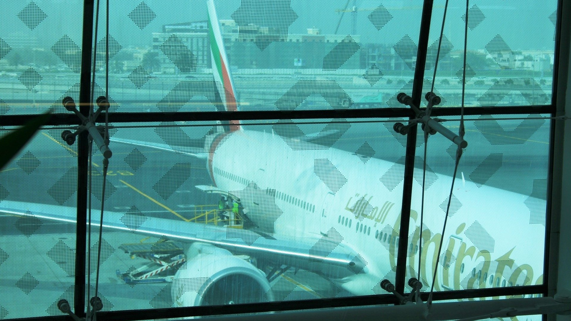 La imagen muestra un avión de la aerolínea Emirates estacionado en un aeropuerto. Se puede ver parte del fuselaje y el ala del avión a través de un gran ventanal con un diseño en las ventanas. Hay trabajadores en la plataforma que parecen estar realizando tareas de mantenimiento o preparación, y se puede observar un entorno aeroportuario con otros edificios y maquinaria en el fondo. La atmósfera es luminosa, aunque un poco difusa debido al cristal.
