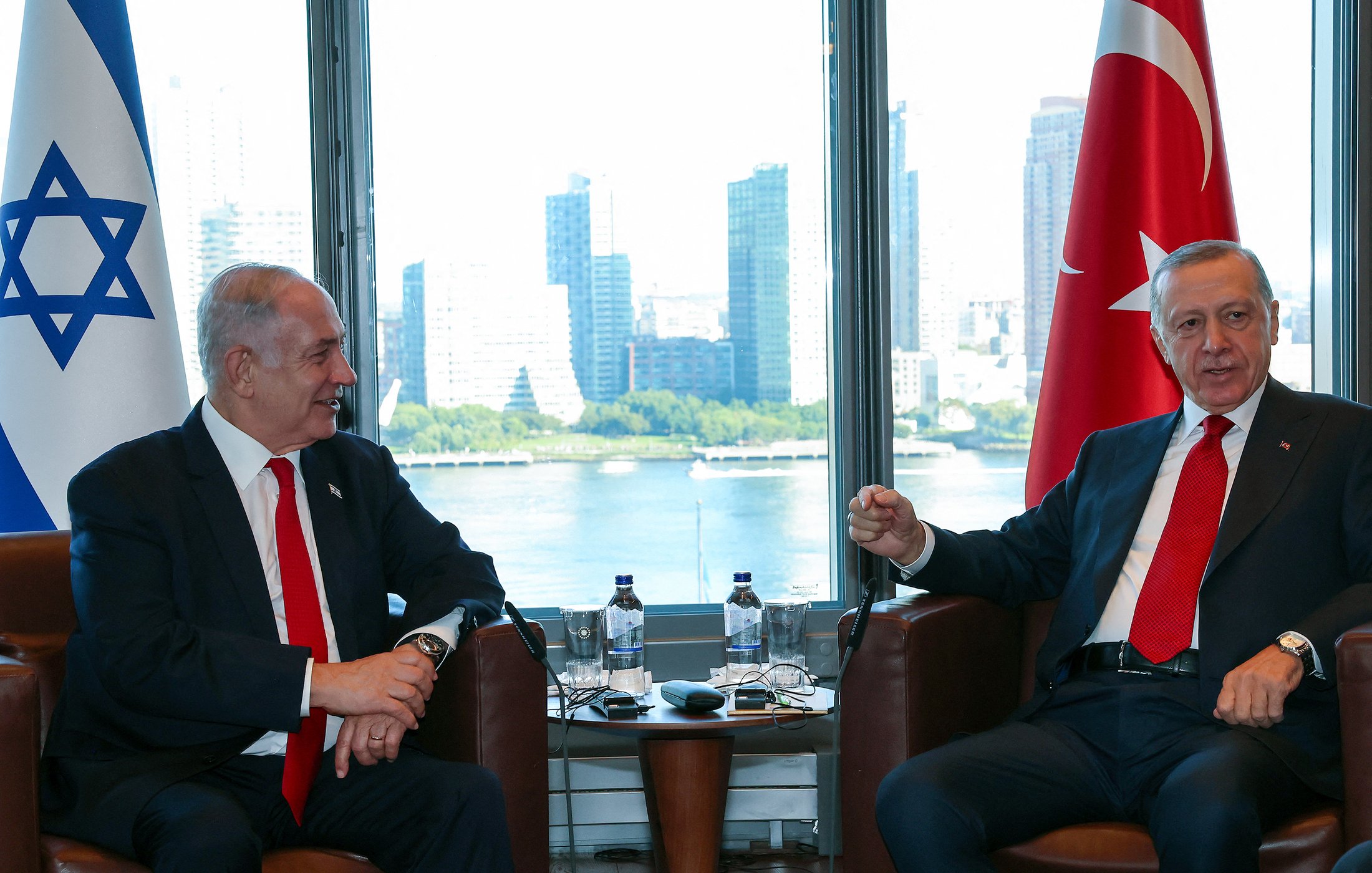 The image shows two men sitting in an office or meeting room with large windows that offer a view of a city skyline in the background. One man appears to be wearing a black suit and a red tie, while the other wears a dark suit and a white shirt with a red tie. There are symbols of Israel and Turkey nearby, indicating a diplomatic or official meeting between representatives of the two countries. The atmosphere seems cordial as they engage in conversation.