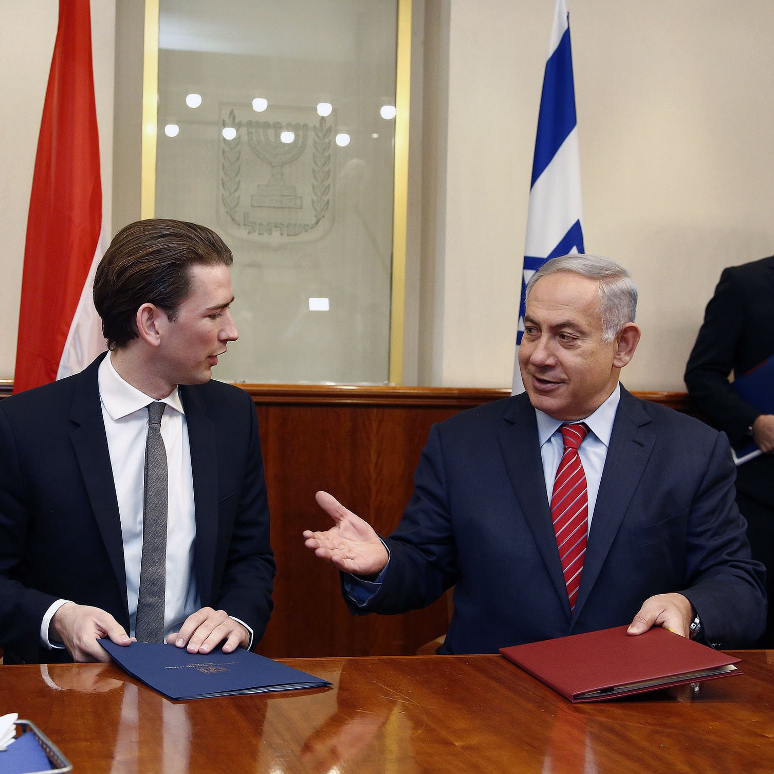 الصورة تظهر مشهداً من اجتماع رسمي، حيث يجلس شخصان على طاولة. الشخص الأول يرتدي بدلة داكنة ولديه شعر قصير، بينما الشخص الثاني يرتدي بدلة مع ربطة عنق حمراء. يبدو أن كلاهما مشارك في نقاش أو اتفاقية رسمية، حيث يوجد خلفهم علمين. الجو العام يبدو جاداً ومهنياً.