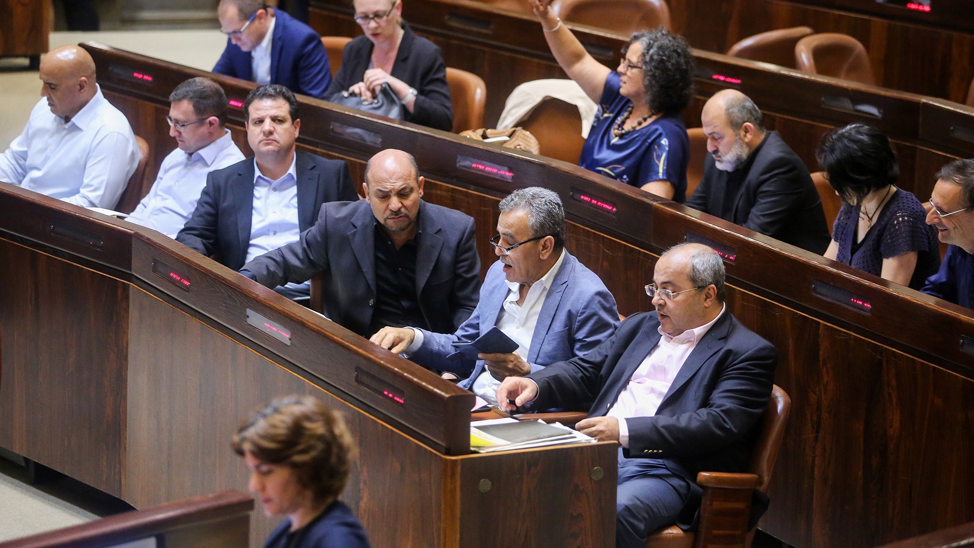 L'image montre une scène à l'intérieur d'une assemblée, probablement dans un parlement. On peut voir plusieurs personnes assises dans des rangées, en train de discuter ou de prendre des notes. Certaines semblent attentives, tandis que d'autres sont engagées dans des conversations. L'environnement est formel, avec des sièges en bois et un éclairage tamisé. Cette composition indique un moment de débat ou de discussion politique.