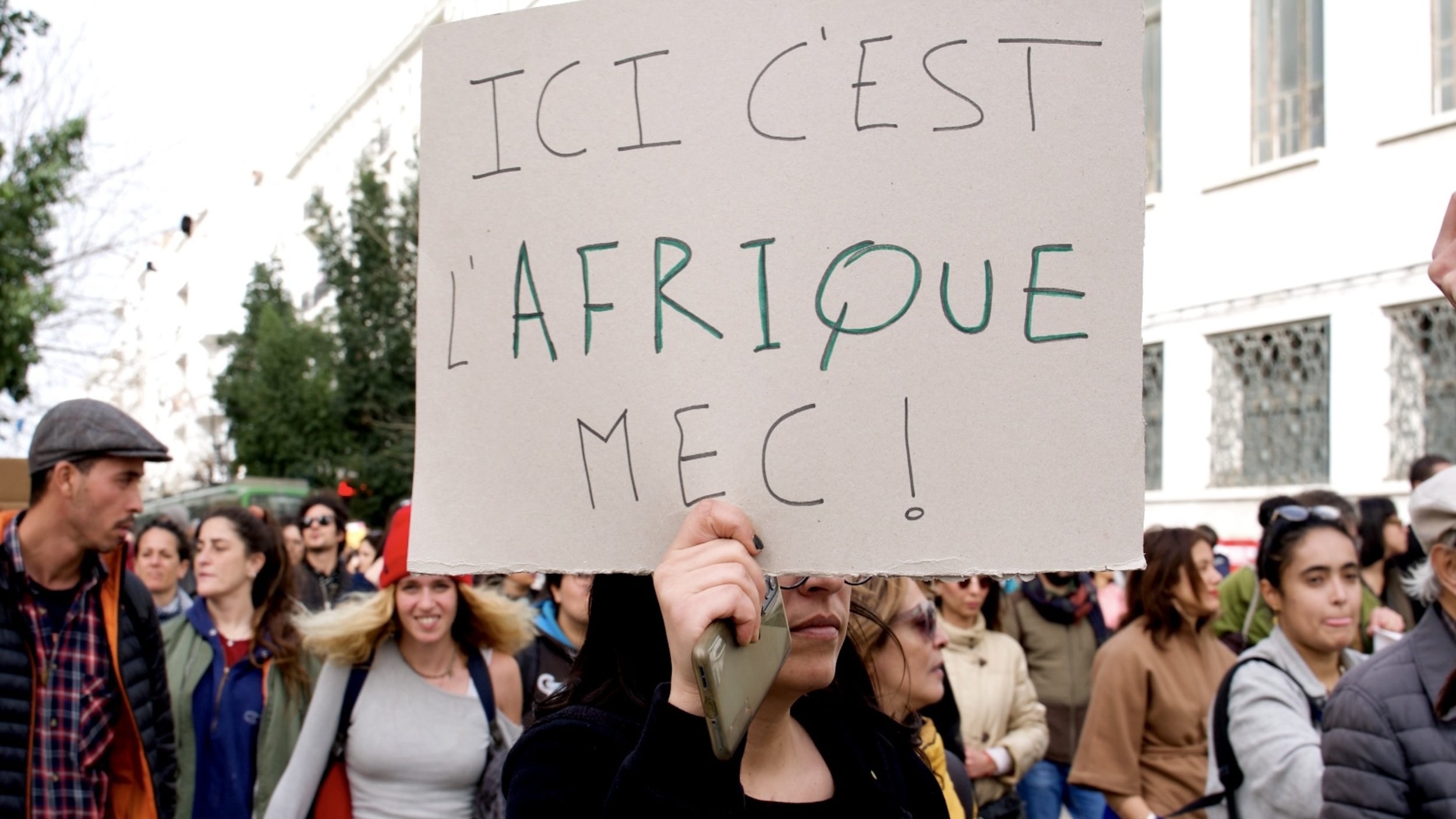 Nell'immagine si vede un gruppo di persone durante una manifestazione. Una donna in primo piano tiene in mano un cartello scritto in francese che recita "ICI C'EST L'AFRIQUE MEC!", il quale esprime un messaggio forte e chiaro. L'atmosfera sembra vivace e partecipativa, con diversi partecipanti che si uniscono per sostenere una causa. Sullo sfondo si notano alberi e edifici, suggerendo un contesto urbano.
