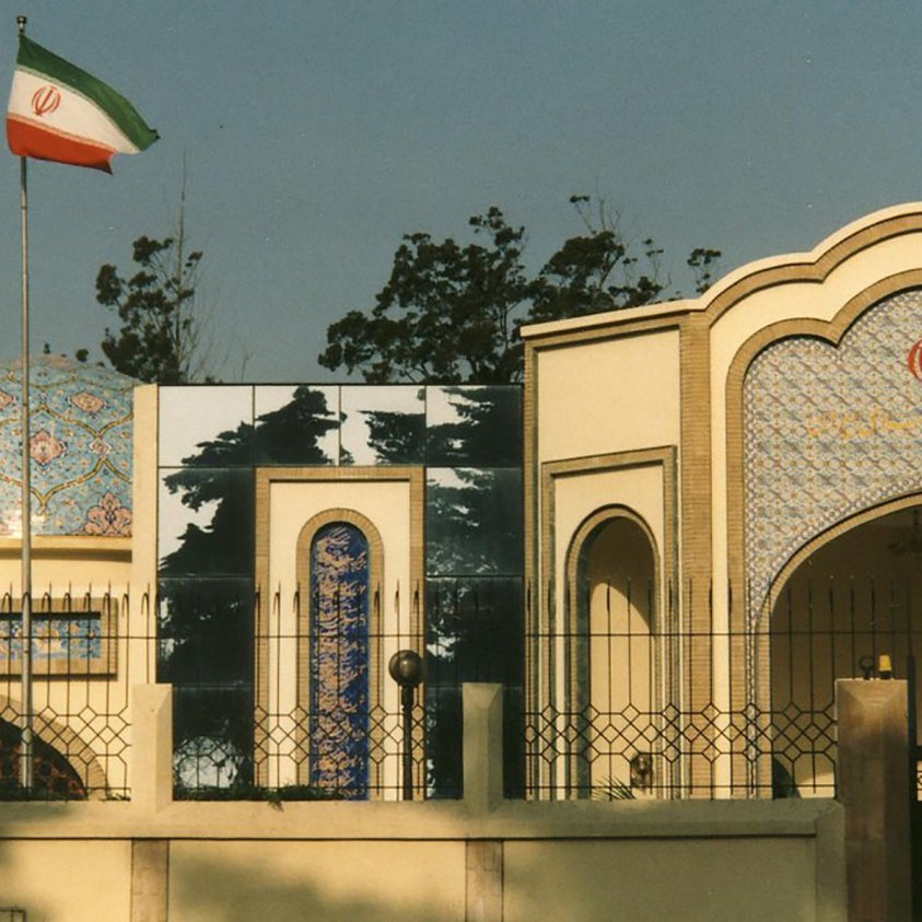 L'image montre un bâtiment architectural de style iranien, typique avec des tuiles colorées et des motifs ornementaux. On peut voir le drapeau iranien flottant sur un mât à gauche. Le bâtiment présente une structure avec un dôme décoré et des arcs, intégrant des éléments de design traditionnels. L'environnement est arboré, avec des arbres visibles en arrière-plan.