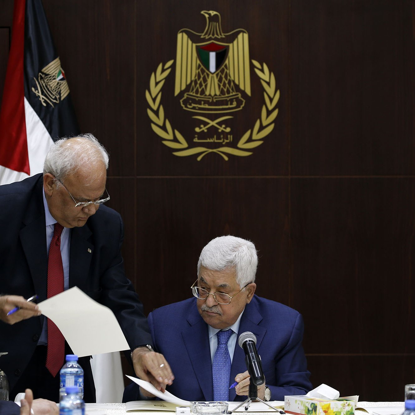 در این تصویر، یک جلسه رسمی در حال برگزاری است. در مرکز تصویر، مردی به نام محمود عباس نشسته است که به نظر می‌رسد در حال مطالعه مدارک یا مستندات است. افراد دیگری نیز در اطراف او نشسته‌اند. یکی از آن‌ها به سمت عباس خم شده و به او مدرکی نشان می‌دهد. در پس‌زمینه، پرچم فلسطین و نماد رسمی مرتبط با دولت فلسطین دیده می‌شود، که به فضایی رسمی و سیاسی اهمیت می‌دهد. میز گردی که آن‌ها دور آن نشسته‌اند، با لوازم اداری و بطری‌های آب تزیین شده است.