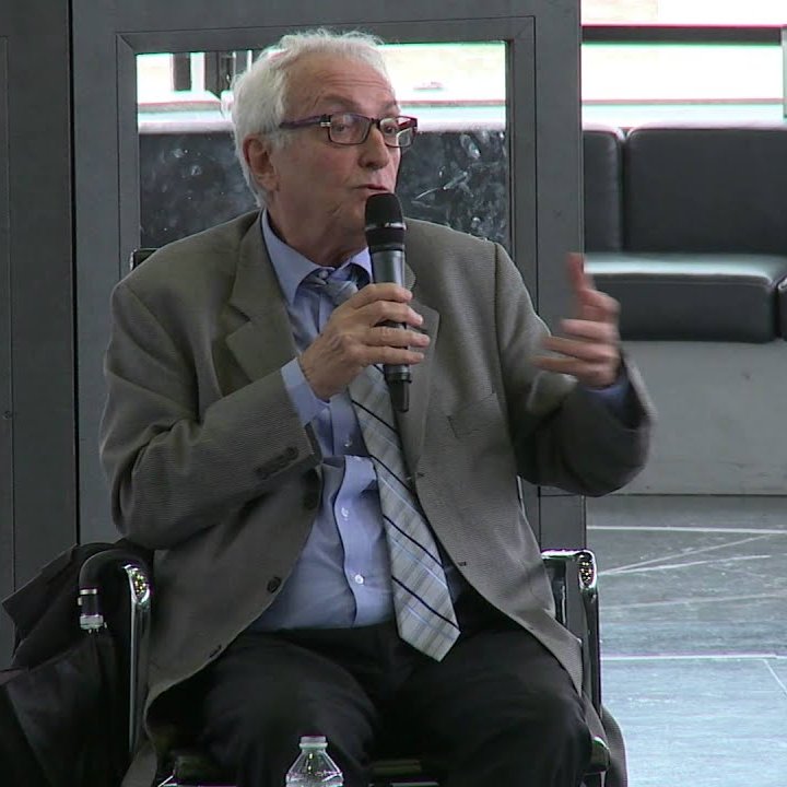 در این تصویر، فردی در حال صحبت کردن با میکروفن است. او کت و شلوار به تن دارد و عینک می‌زند. به نظر می‌رسد در یک جلسه یا کنفرانس شرکت کرده و به حاضران ارائه‌ای می‌دهد. زمینه تصویر نشان‌دهنده یک فضای مدرن و باز است.