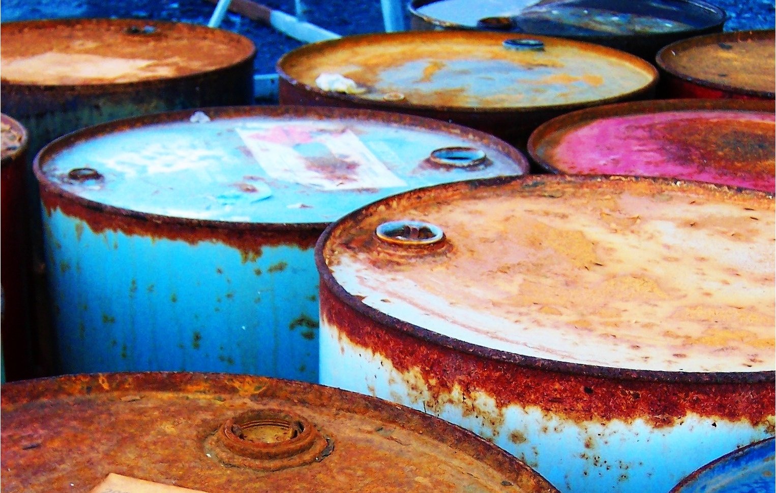 L'image montre plusieurs fûts en métal, probablement des fûts de pétrole ou d'huile. Ils sont empilés les uns à côté des autres, avec des couvercles rouillés et des étiquettes décolorées. La surface des fûts présente des signes d'oxydation, ce qui leur donne un aspect usé et ancien. Les couleurs variées du métal, allant du rouille au bleu clair, ajoutent à l'esthétique générale de la scène.