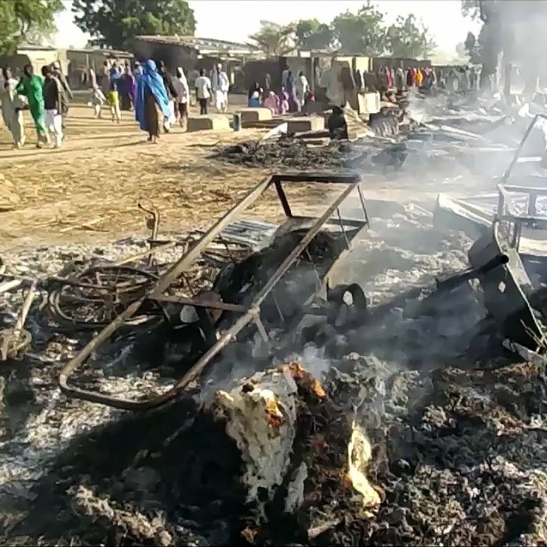 La imagen muestra un escenario devastador después de un incendio. Hay estructuras de metal chamuscadas y escombros humeantes en el suelo. En el fondo, algunas personas parecen estar observando la escena, mientras que el humo se eleva en el aire. El paisaje refleja una atmósfera de destrucción, posiblemente en un área rural o un mercado.