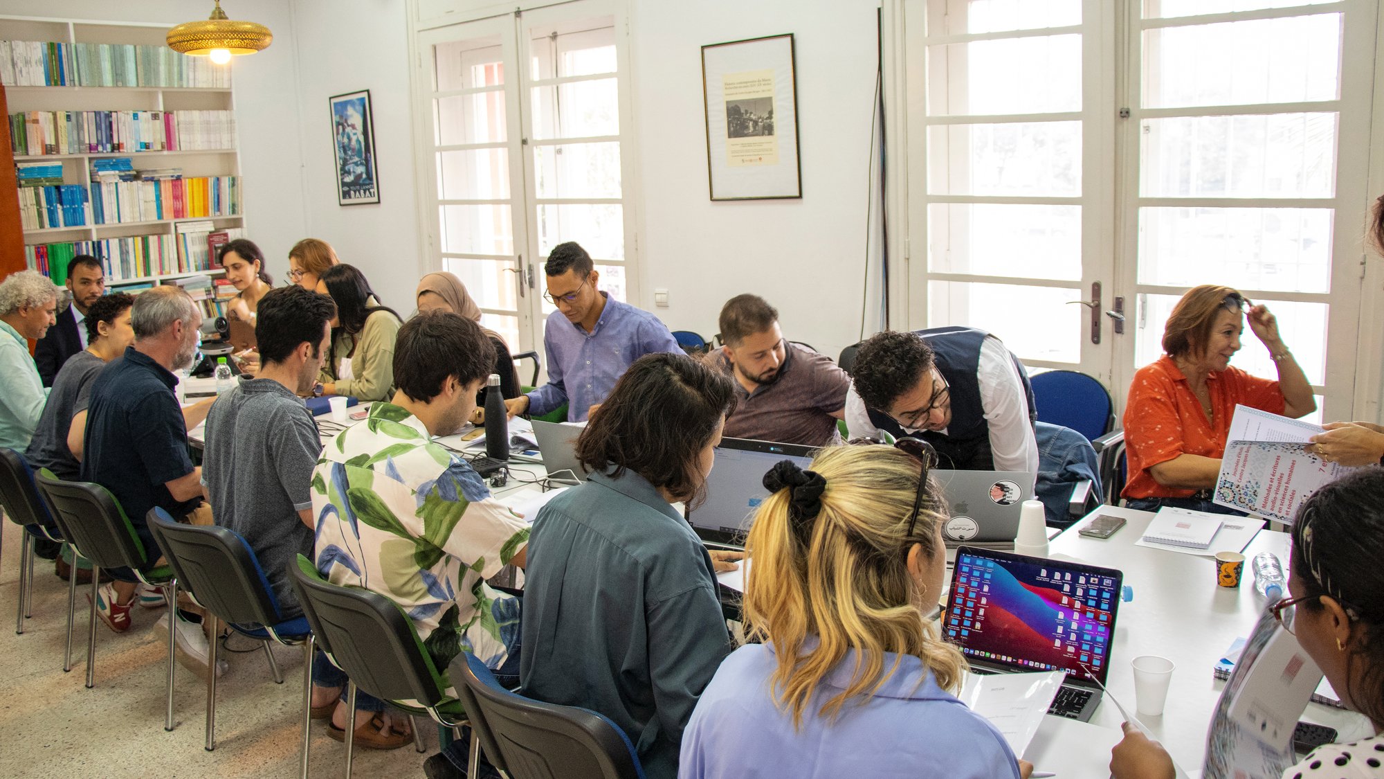 L'image montre une salle de réunion où un groupe de personnes est assis autour d'une grande table. Plusieurs participants semblent travailler sur des ordinateurs portables, tandis que d'autres consultent des documents. Les murs sont décorés de livres et d'illustrations, et plusieurs fenêtres laissent entrer la lumière naturelle. L'ambiance apparaît studieuse et collaborative.