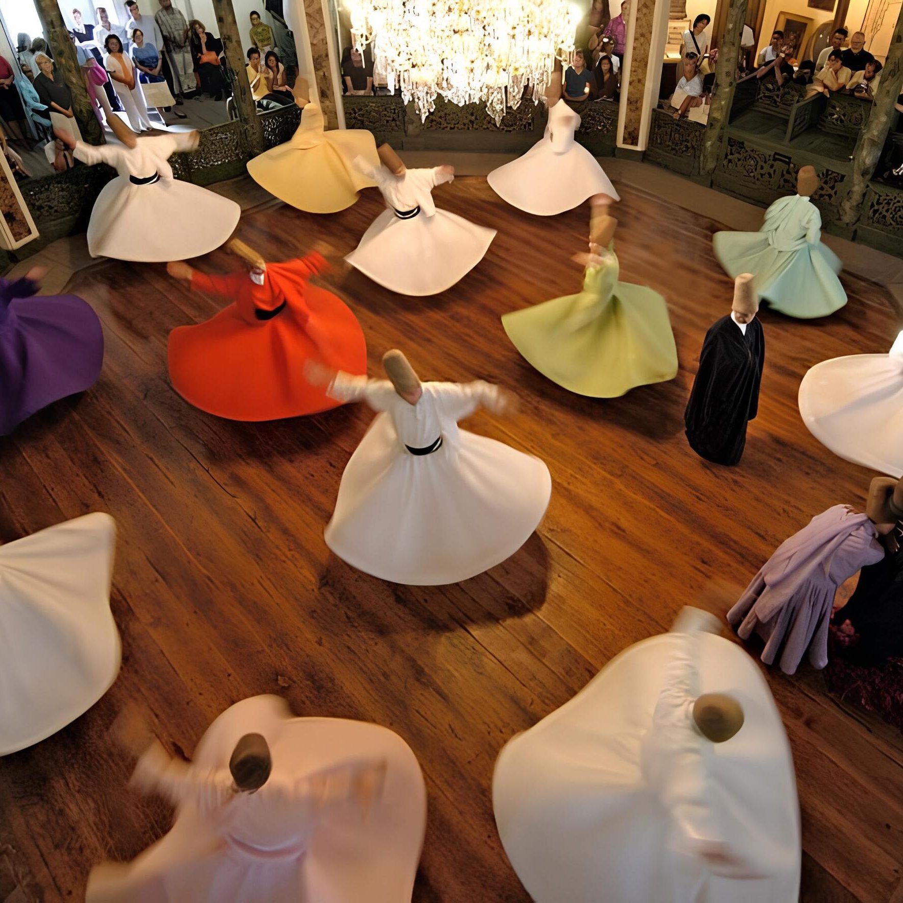 L'immagine mostra un gruppo di ballerini che si esibiscono in una danza tradizionale, con abiti ampi e colorati che girano in cerchio. I ballerini sono disposti in un grande spazio circolare, con un pavimento in legno lucido e una decorazione elegante attorno a loro. Al centro della scena, un lampadario di cristallo illumina l'ambiente, mentre il pubblico osserva l'esibizione con interesse. I colori degli abiti, che vanno dal bianco al viola e arancio, creano un'atmosfera vibrante e festosa.