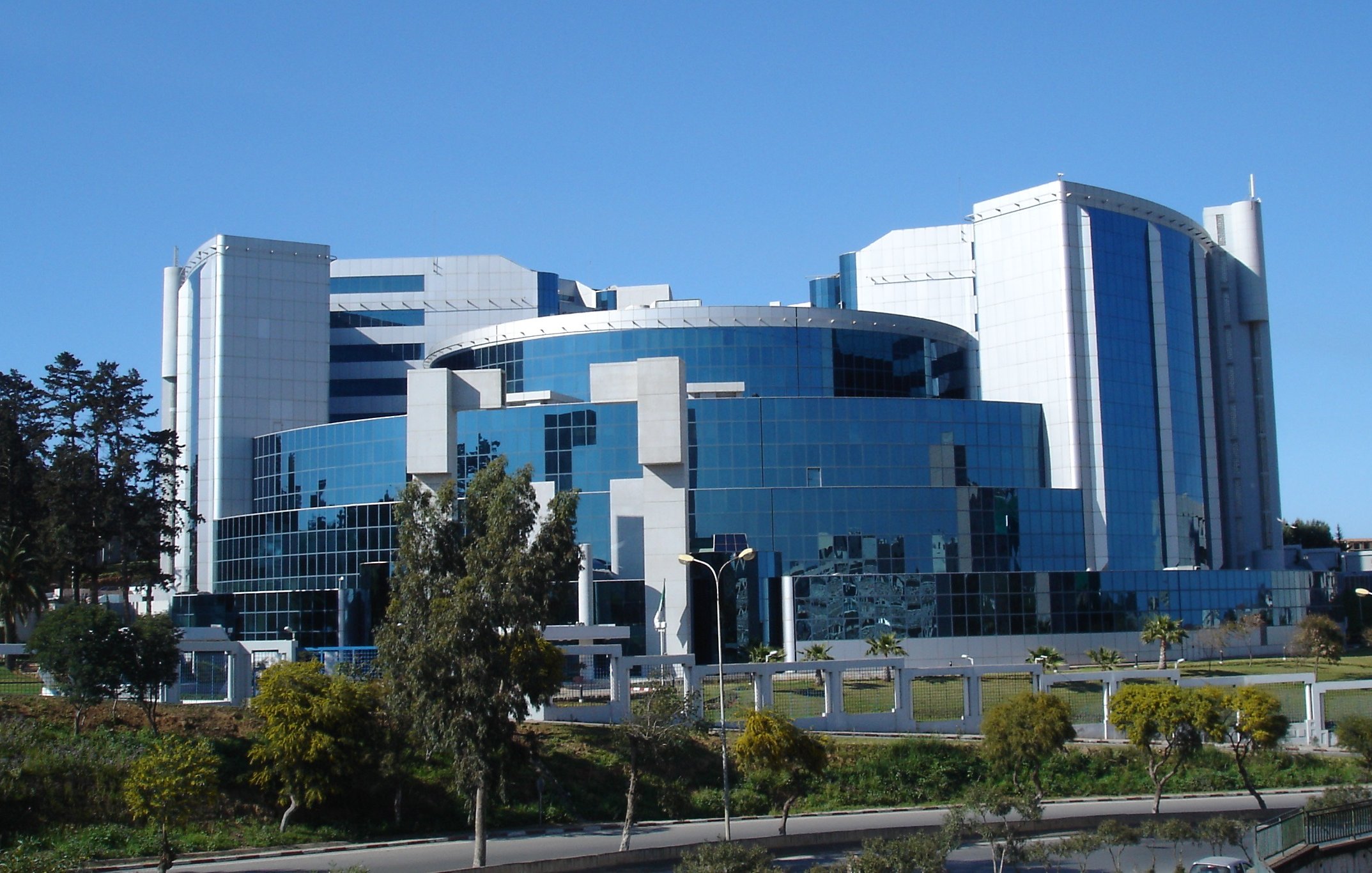 L'image montre un bâtiment moderne avec une architecture contemporaine caractérisée par des façades en verre et des formes géométriques. Le bâtiment semble être de grande taille, avec plusieurs niveaux et une combinaison de matériaux tels que le métal et le verre. On peut également voir quelques arbres et végétation autour, ainsi qu'un ciel bleu clair en arrière-plan. L'ensemble dégage une impression de modernité et de sophistication.
