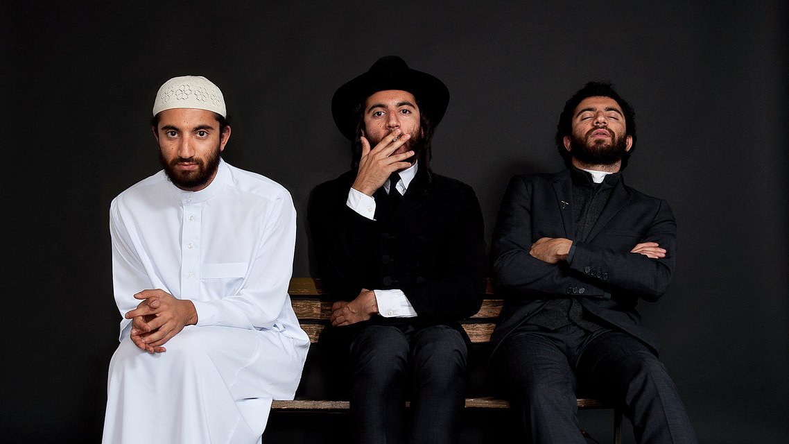 L'image montre trois hommes assis sur un banc, chacun portant un style vestimentaire distinct. À gauche, un homme en blanc, portant une tenue traditionnelle et un couvre-chef. Au centre, un homme habillé de manière formelle, avec un chapeau noir et une tenue sombre. À droite, un homme également en costume, avec une expression pensive. Le fond est noir, ce qui met en valeur les vêtements et les postures des sujets.