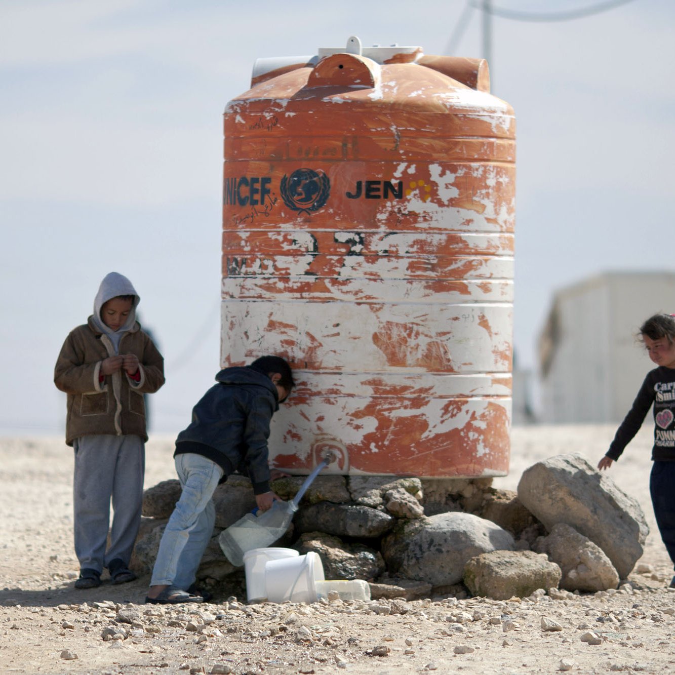 L'image montre un groupe d'enfants autour d'un réservoir d'eau. Le réservoir, de couleur orange et marqué par des signes d'usure, semble abriter l'eau potable. Les enfants, dont l'un est en train de remplir un récipient, sont habillés de manière simple et semblent se trouver dans un environnement désertique. On peut voir des pierres autour du réservoir, et le paysage en arrière-plan est sobre, suggérant une situation de besoin en ressources.
