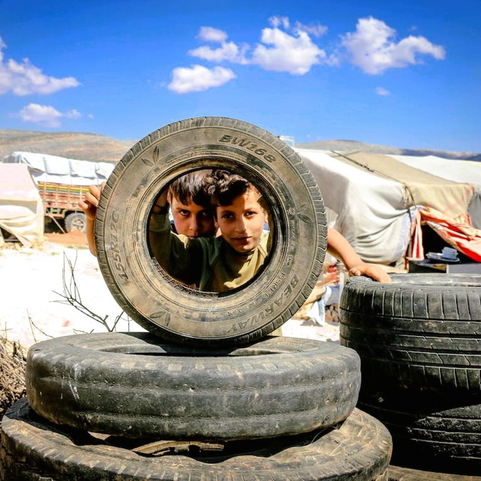 در این تصویر، دو کودک در حال نگاه کردن از میان یک لاستیک بزرگ هستند. لاستیک‌ها به صورت عمومی به عنوان بخشی از یک ساخت و ساز یا بازی در محوطه‌ای از فضای باز قرار داده شده‌اند. پس‌زمینه شامل چادرها و مناظر طبیعی است. آسمان آبی و ابرهای پراکنده در بالای سر دیده می‌شوند. فضای کلی تصویر حس کنجکاوی و بازی کودکانه را منتقل می‌کند.