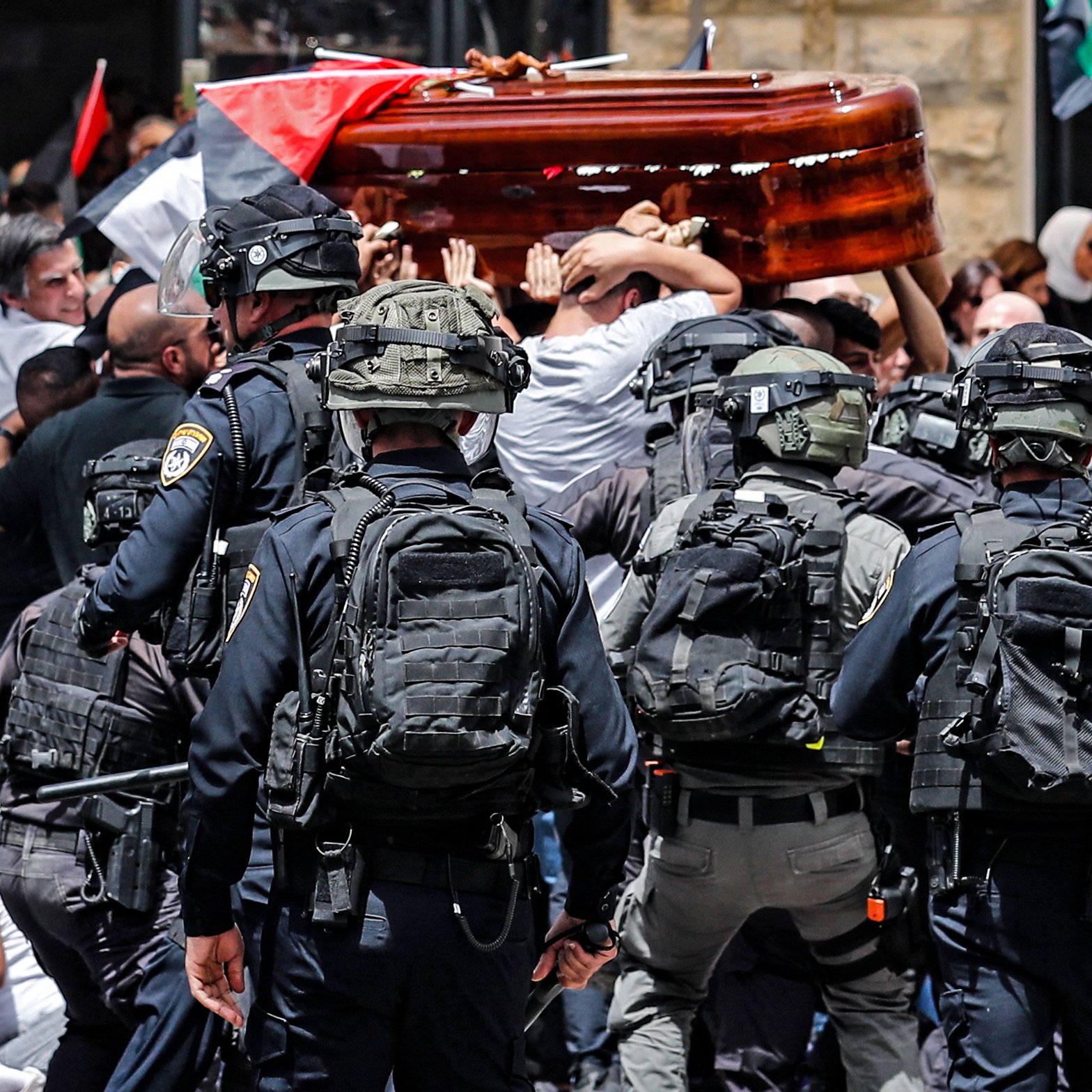 تظهر الصورة مشهدًا من حدث عام حيث يحدث تداخل بين مجموعة من الناس وقوات الأمن. يتم حمل تابوت خشبي من قبل المشاركين في الهتافات، بينما يحيط بهم رجال شرطة يرتدون تجهيزات خاصة. الخلفية تضم أعلامًا، مما يشير إلى أن هذه الفعالية قد تكون مرتبطة بمسألة وطنية أو سياسية. الجو يبدو مشحونًا بالانفعالات، مع وجود حشد من الناس في حالة من التوتر.