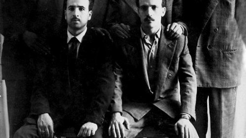 L'image montre un groupe de six hommes, habillés en costumes, posant ensemble. Ils sont disposés sur deux niveaux : trois debout à l'arrière et trois assis à l'avant. Tous les hommes affichent une expression sérieuse et se tiennent près les uns des autres, ce qui donne une impression d'unité. L'arrière-plan est neutre, avec des rideaux qui ajoutent une texture à la scène. La photo semble être en noir et blanc, ce qui lui confère un aspect vintage.