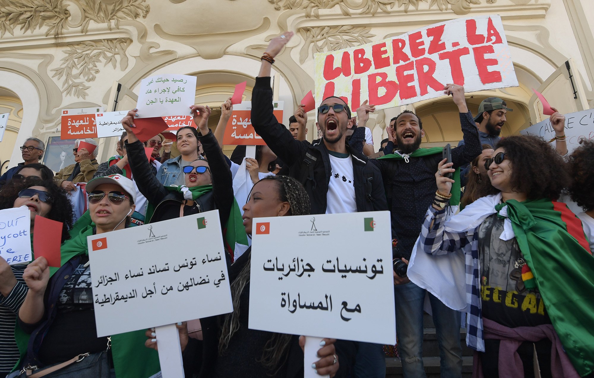L'image montre un groupe de personnes rassemblées lors d'une manifestation. Les manifestants portent des pancartes et des drapeaux, exprimant des slogans tels que "Libérez la liberté". Les participants semblent engagés et passionnés, brandissant des affiches avec des messages en arabe et en français, appelant à la justice et à l'égalité. L'ambiance est énergique et déterminée, témoignant d'une dynamique de revendication sociale ou politique. L'arrière-plan présente une architecture caractéristique, ajoutant au contexte de l'événement.