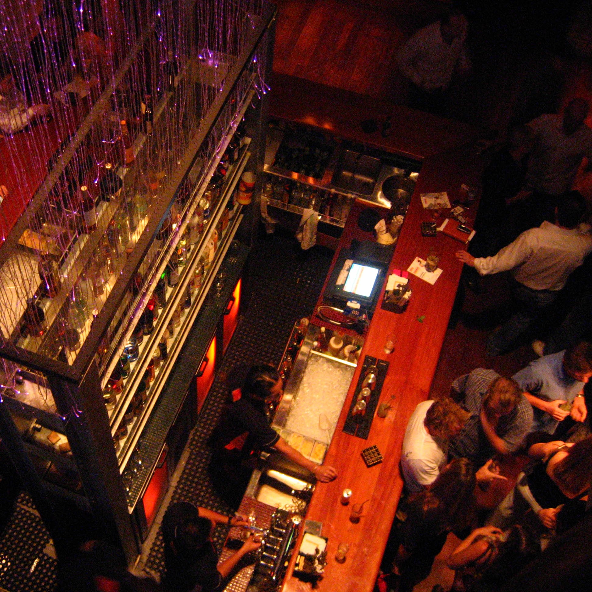 The image depicts a lively bar scene viewed from above. The bar features multiple shelves filled with bottles and is adorned with decorative lights. People are gathered throughout the space, engaging in conversation and enjoying drinks. The atmosphere seems vibrant and social, with a mix of patrons at the bar and tables. The lighting adds a warm, inviting ambiance to the setting.