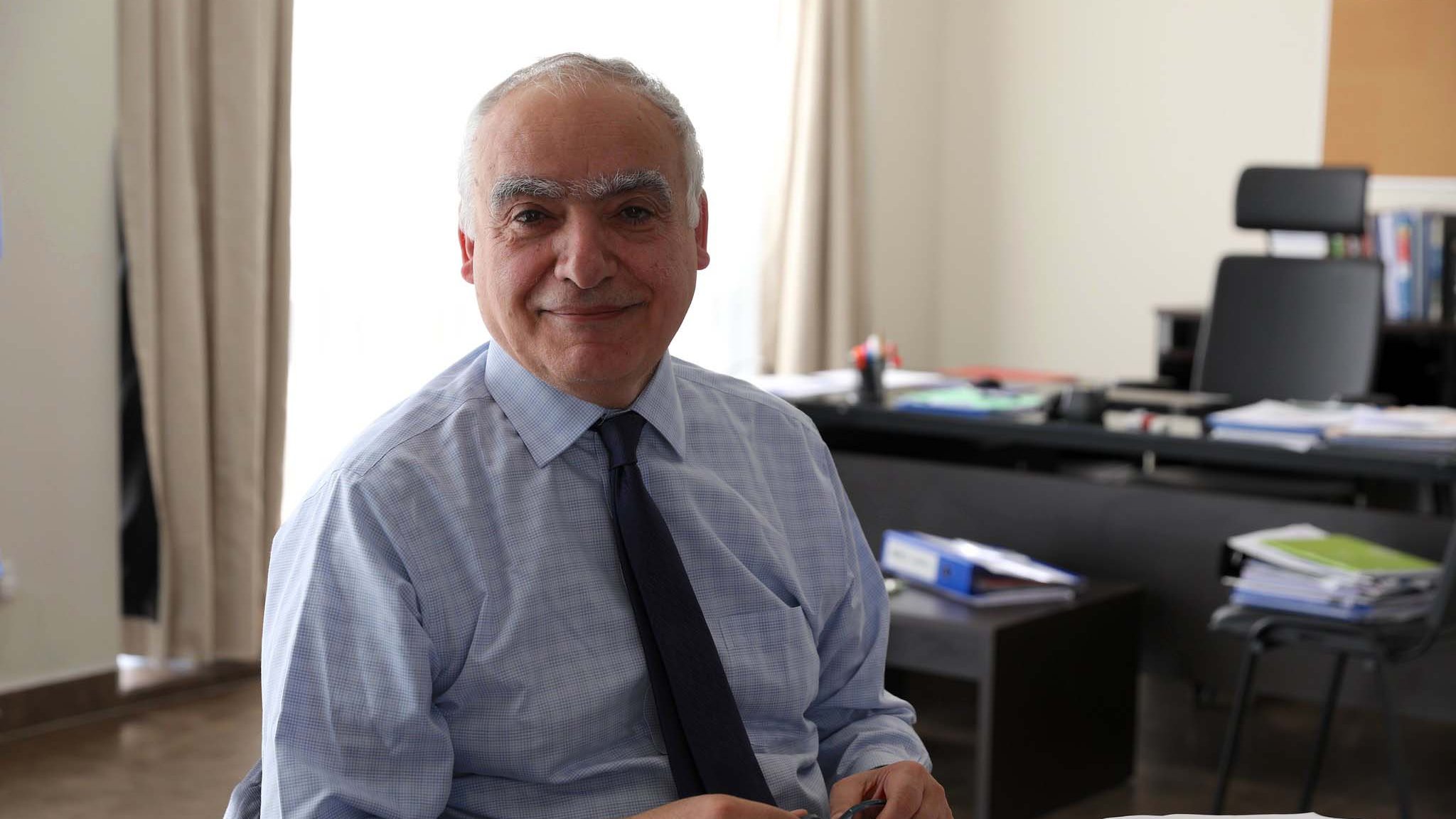 L'immagine mostra un uomo sorridente seduto a un tavolo in un ambiente di lavoro. Indossa una camicia e una cravatta, e sembra essere concentrato su alcune carte e documenti posti davanti a lui. Sullo sfondo, si intravedono elementi di un ufficio, come una scrivania con vari materiali e un ambiente luminoso grazie alla luce naturale che entra dalla finestra.