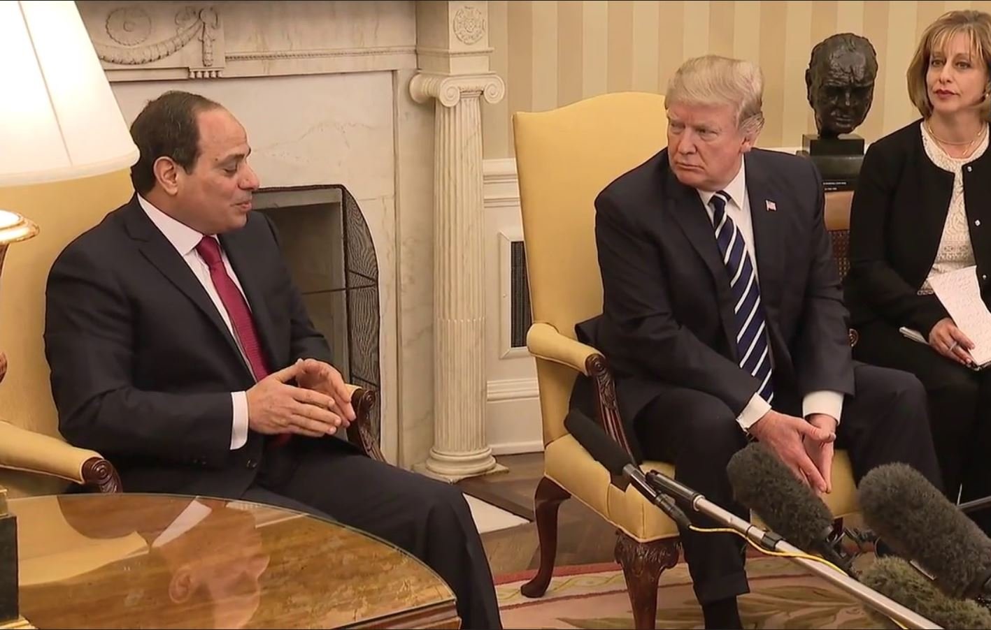 The image shows two men sitting in a formal setting, likely a meeting room in a government building. One man, on the left, is wearing a dark suit and a red tie, appearing to speak or make a point. The other man, on the right, is dressed in a dark suit with a striped tie and is attentively listening or observing. There are microphones positioned between them, indicating that their conversation is being recorded or broadcast. The background features a light-colored wall and a decorative element, possibly a bust or sculpture, along with a warm lamp on the table.