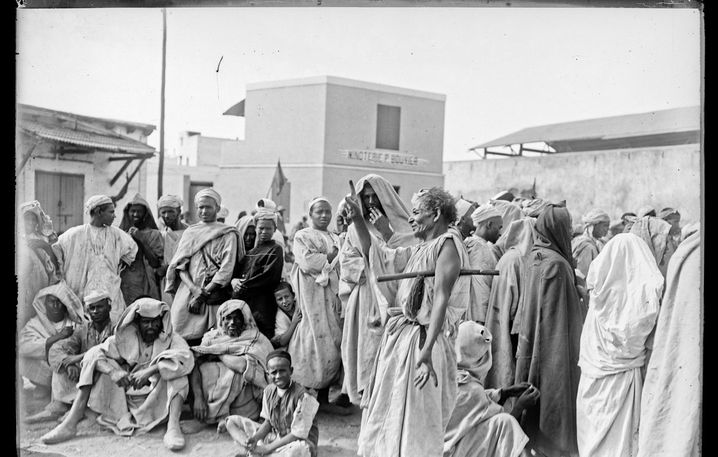 تظهر الصورة مشهداً تاريخياً يضم مجموعة من الأشخاص يرتدون ملابس تقليدية في بيئة حضرية. يتواجد الرجال والنساء بشكل مختلط، بعضهم يجلس وآخرون واقفون، حيث يبدو أنهم يعيشون لحظة تتسم بالنقاش أو الاحتجاج. الخلفية تضم مباني بسيطة، مما يشير إلى بيئة قد تكون في منطقة شمال إفريقيا أو الشرق الأوسط. تعكس الملابس والتعبيرات الثقافية تفاصيل الحياة الاجتماعية في تلك الفترة.