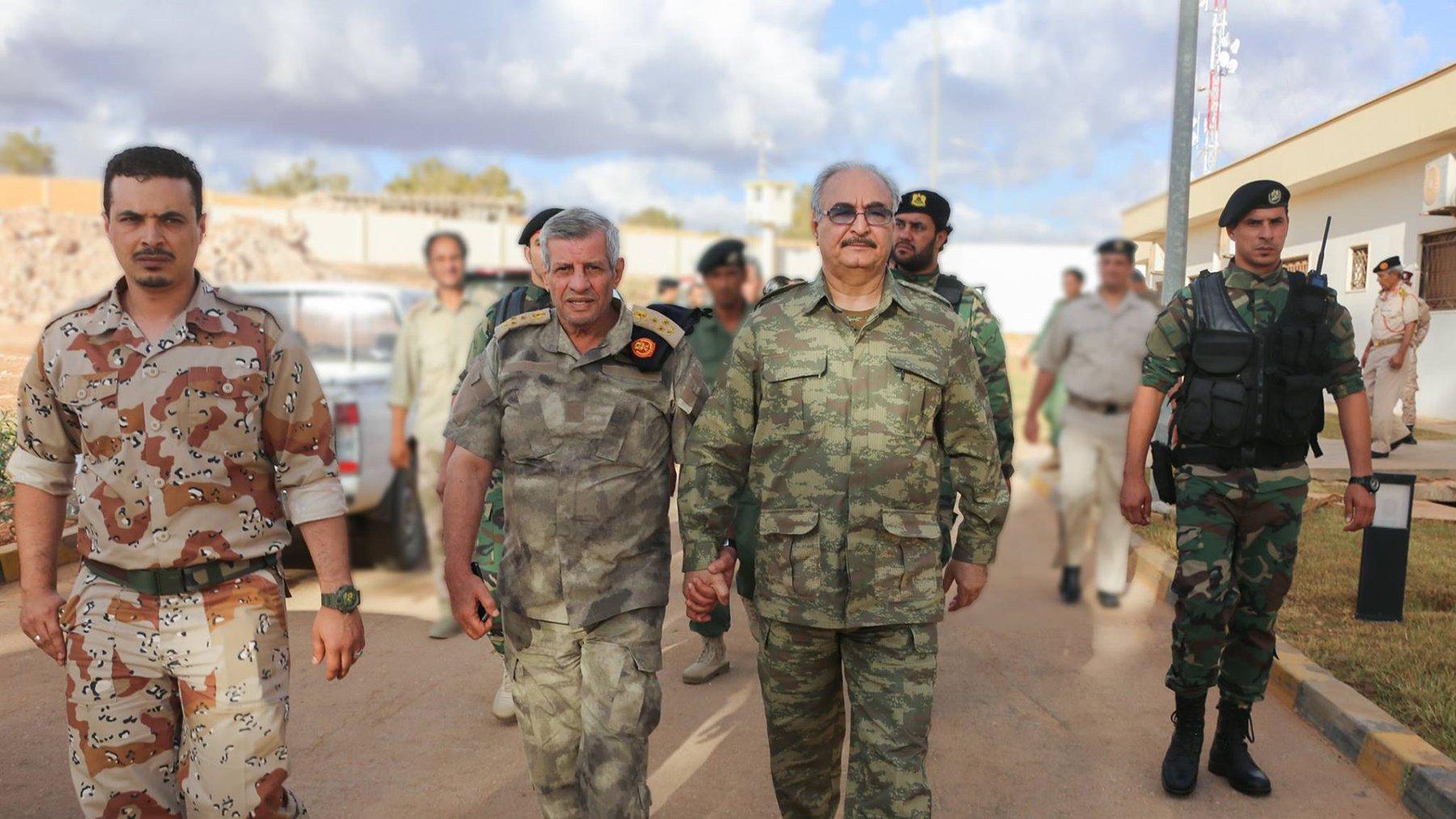 The image depicts a group of military personnel walking together. Two men in the foreground, wearing military uniforms, are holding hands and appear to be engaged in conversation. One is in a camouflage uniform, while the other has a distinctive pattern. They are flanked by soldiers in various military attire in the background. The setting appears to be an outdoor area, possibly a military base, with vehicles and other personnel visible. The sky shows some clouds, indicating daytime.
