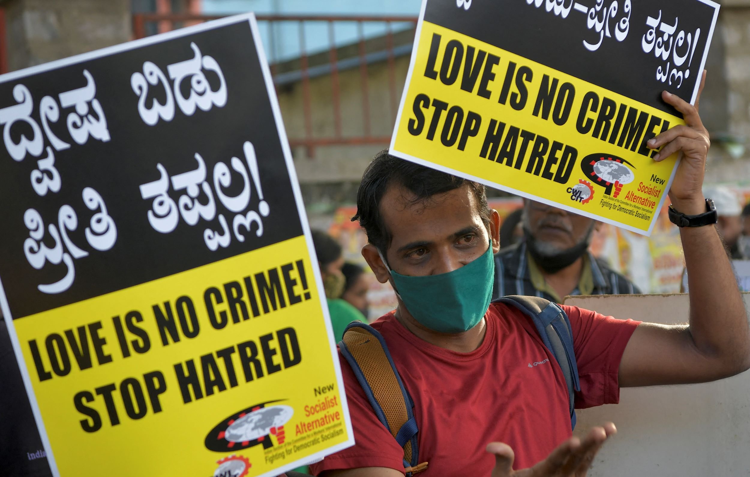 The image depicts a person holding two large signs that read "LOVE IS NO CRIME! STOP HATRED!" in both English and Kannada, a language spoken in India. The individual is wearing a face mask and appears to be participating in a demonstration or rally, advocating for love and against hate. The atmosphere suggests a sense of activism and a call for social change, with a crowd visible in the background. The signs are bright and eye-catching, emphasizing the message strongly.