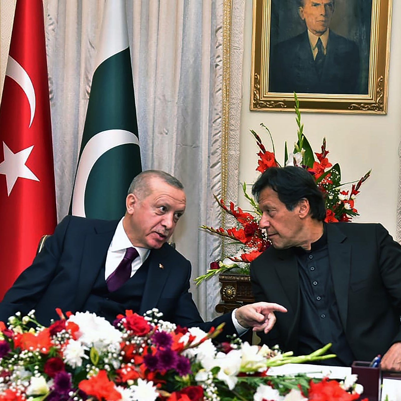 The image features two men engaged in conversation, likely discussing matters of significance. They are seated at a table adorned with flowers, and the backdrop includes flags of Turkey and Pakistan prominently displayed. A portrait of a historical figure hangs on the wall above them. Both individuals appear to be in formal attire, suggesting an official or diplomatic context for their meeting.
