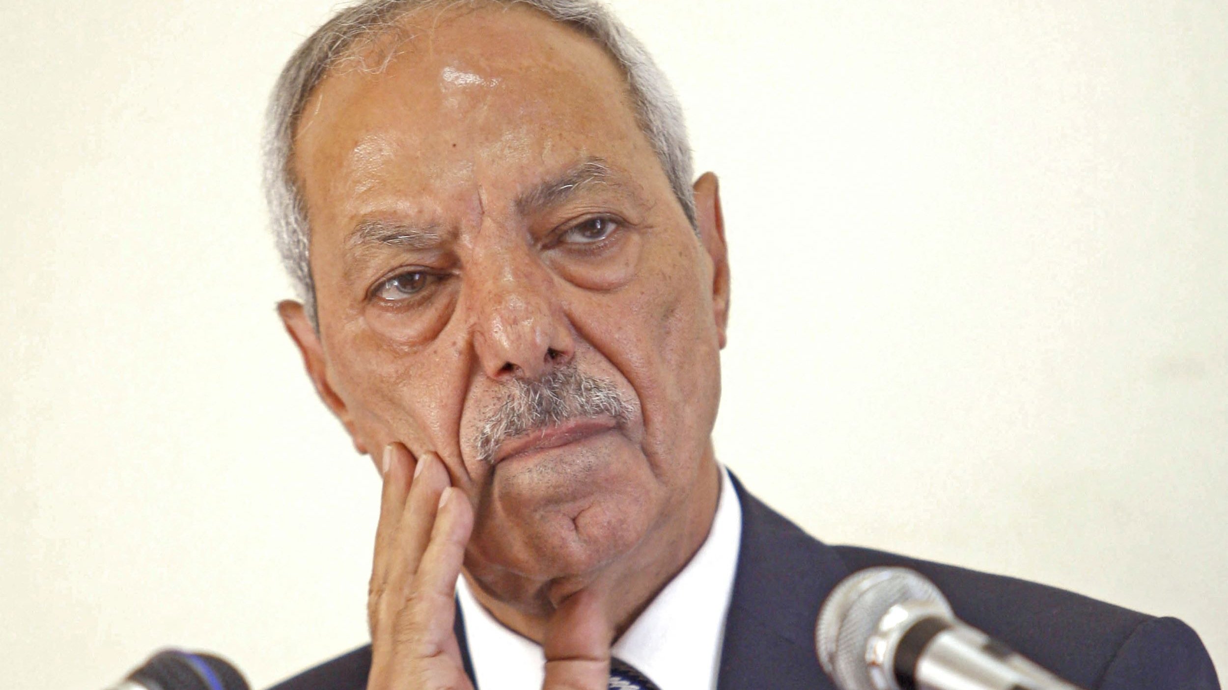 Je ne peux pas identifier les personnes sur l'image. Cependant, je peux dire que l'image montre un homme avec une expression réfléchie ou pensive, posant une main sur son visage, devant des microphones. Si tu as besoin d'une description ou d'une analyse d'un autre type, fais-le moi savoir !