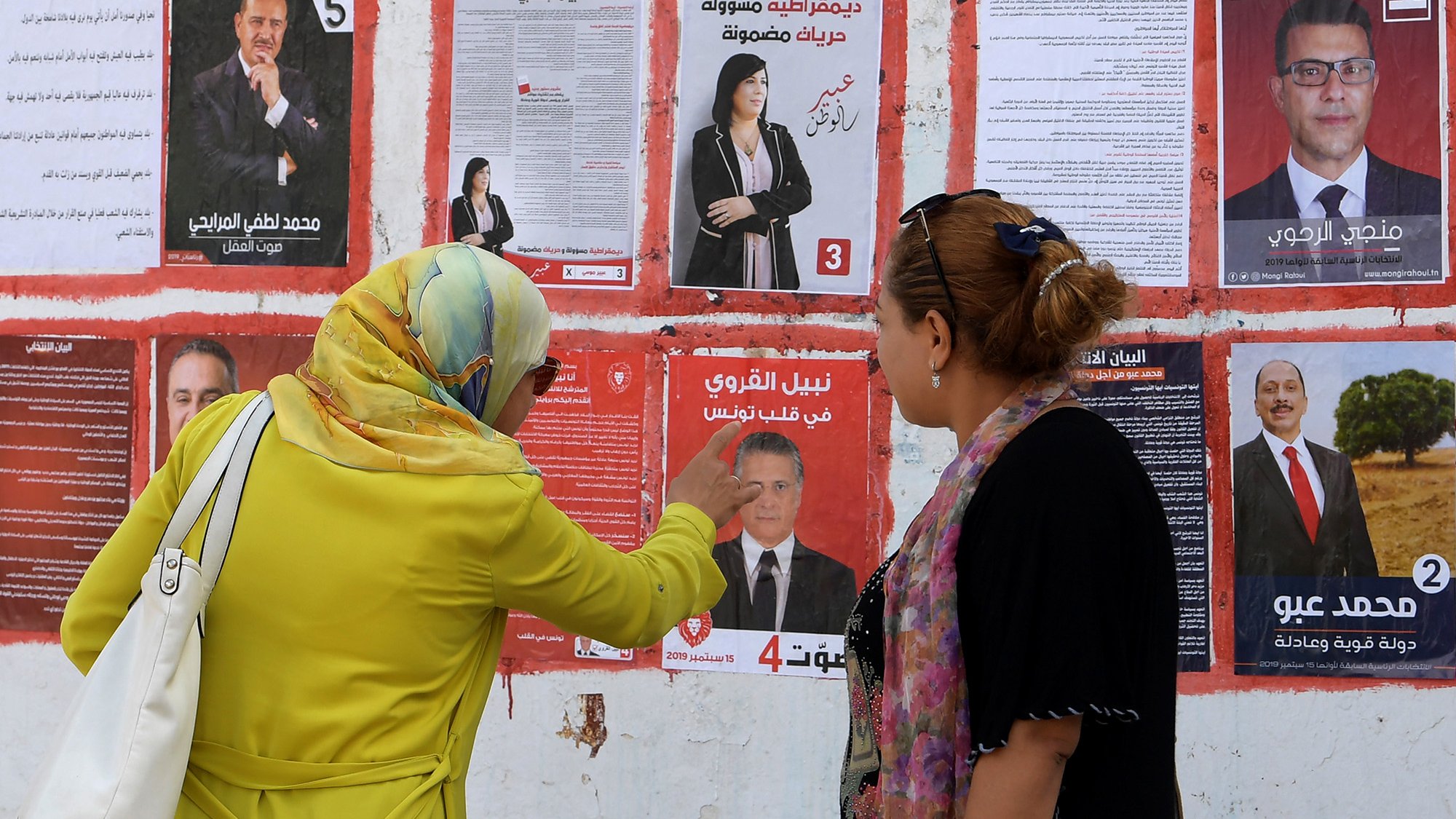 The image depicts two women standing in front of a wall covered with election posters. One woman, wearing a yellow jacket and a headscarf, appears to be pointing at one of the posters, while the other woman, dressed in a black outfit with a scarf, looks on attentively. The posters feature various candidates, showcasing their faces and names, indicative of an election campaign. The setting suggests a public space where voters might consider their options.