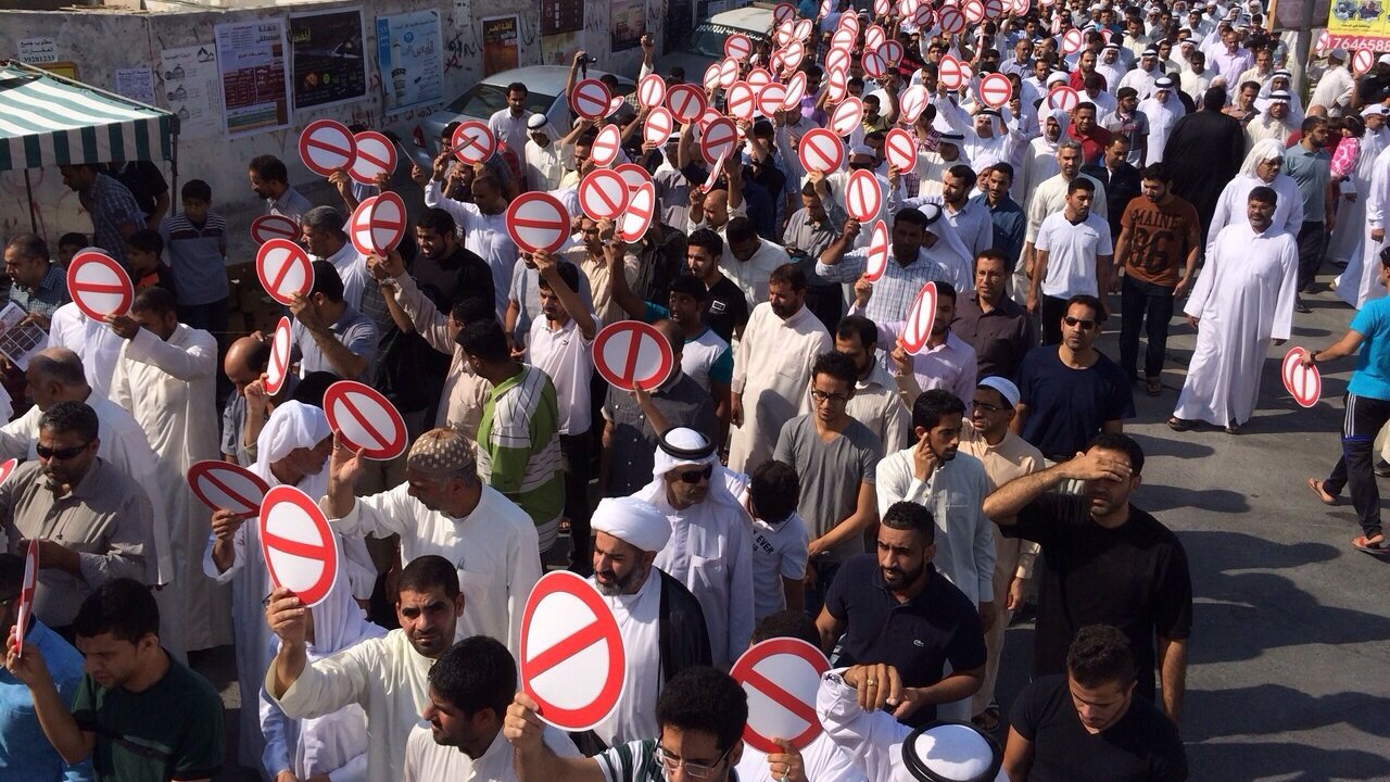L'image montre une grande foule de personnes rassemblées dans une rue. Les manifestants brandissent des pancartes avec un symbole de cercle rouge barré, souvent utilisé pour signifier une interdiction. Les participants portent des vêtements variés, certains en tenue traditionnelle. On observe des hommes et des femmes de différentes origines, tous unis dans leur démarche. L'environnement urbain en arrière-plan, avec des bâtiments et des affiches, suggère un contexte de protestation ou de manifestation.