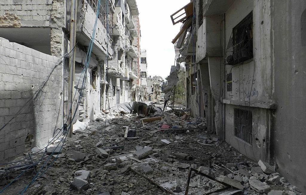L'image montre une rue dévastée, probablement dans une zone de conflit. Les bâtiments de chaque côté sont endommagés, avec des murs effondrés et des débris éparpillés sur le sol. Des fils électriques pendent, et l'atmosphère générale dégage une impression de destruction et d'abandon. On peut observer des traces des conséquences des affrontements, avec un paysage urbain sévèrement altéré.