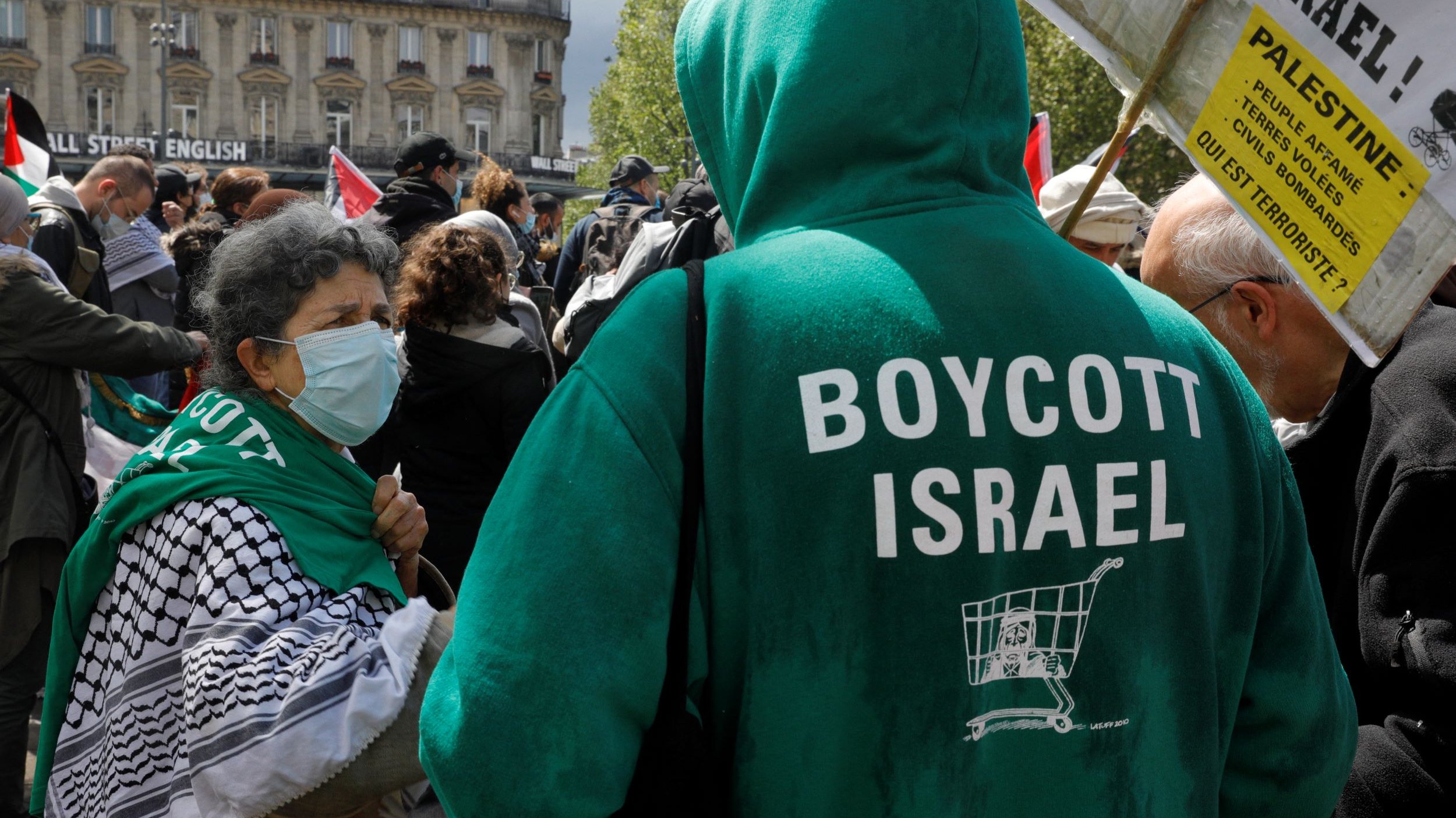 The image depicts a gathering or protest where individuals are expressing their political views. In the foreground, a person wearing a green hoodie that reads "BOYCOTT ISRAEL" is facing another individual. The latter is wearing a face mask and has a traditional black-and-white patterned scarf. The setting appears to be in a public space, with banners and flags present in the background, indicating a demonstration related to Palestinian rights or opposition to certain policies regarding Israel.