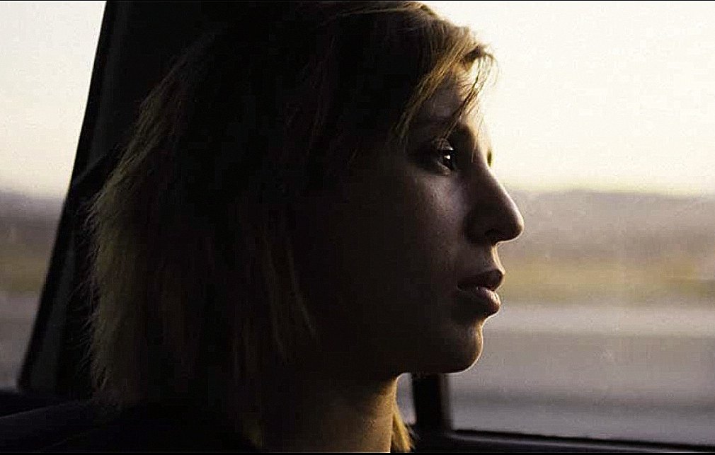 L'image montre une silhouette d'une personne assise à l'intérieur d'une voiture. Elle regarde par la fenêtre, prenant un moment de réflexion. La lumière est douce, créant une ambiance mélancolique, et le paysage flou en arrière-plan suggère un voyage en cours. Le visage de la personne est partiellement éclairé, mettant en valeur une expression pensante.