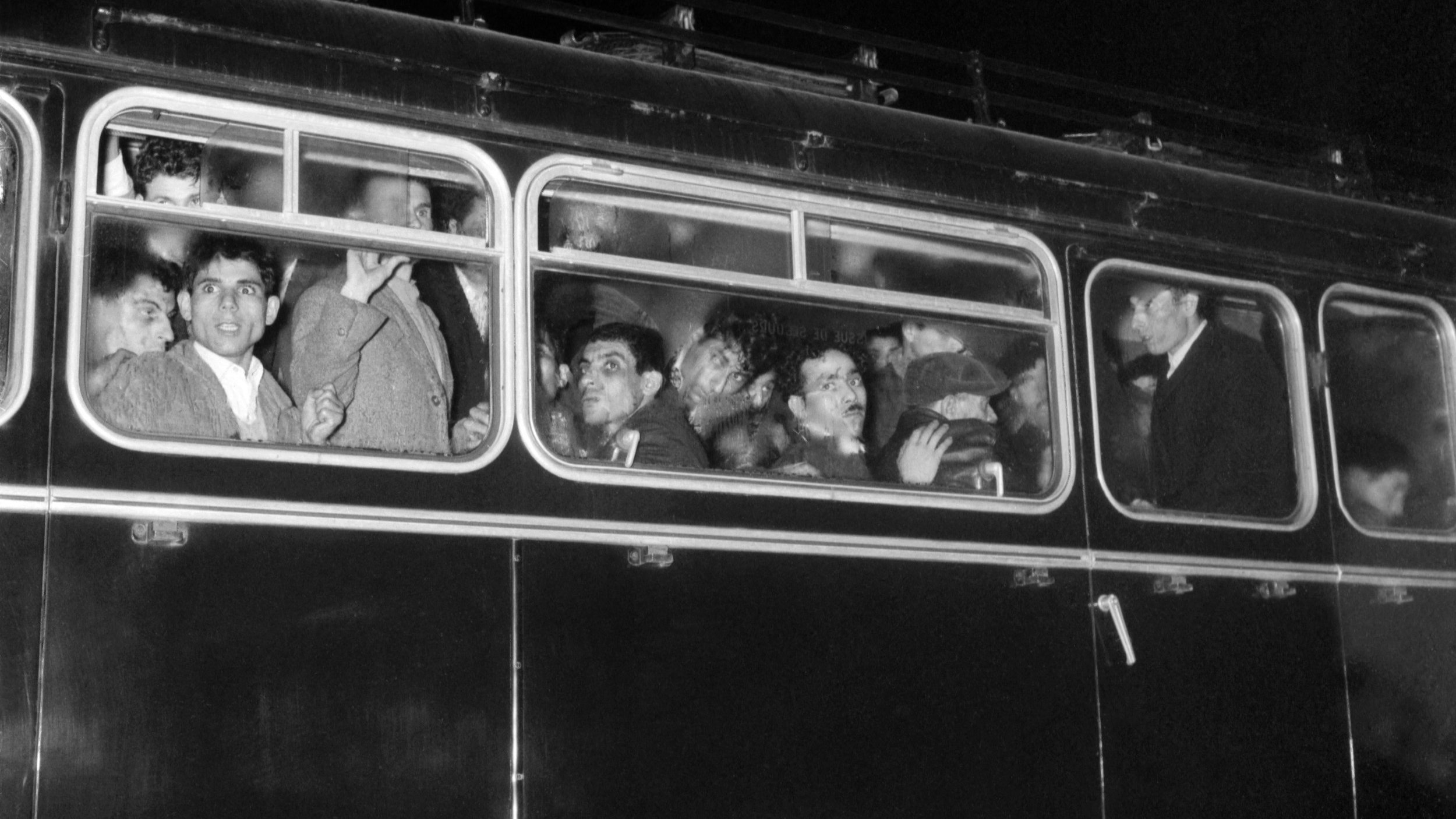 تظهر الصورة حافلة قديمة في الليل، وهي مزدحمة بالركاب الذين يبدون في حالة من الحماس أو الانفعالات. بعض الأشخاص ينظرون من النوافذ في اتجاه الكاميرا، بينما يبدو الآخرون مشغولين بالحديث أو التفاعل مع بعضهم. الأضواء الخافتة تضفي جواً مميزاً على المشهد، مما يعكس لحظة مميزة أو حدثاً معيناً. الحافلة تتميز بتصميمها الكلاسيكي، مما يشير إلى فترة زمنية سابقة.