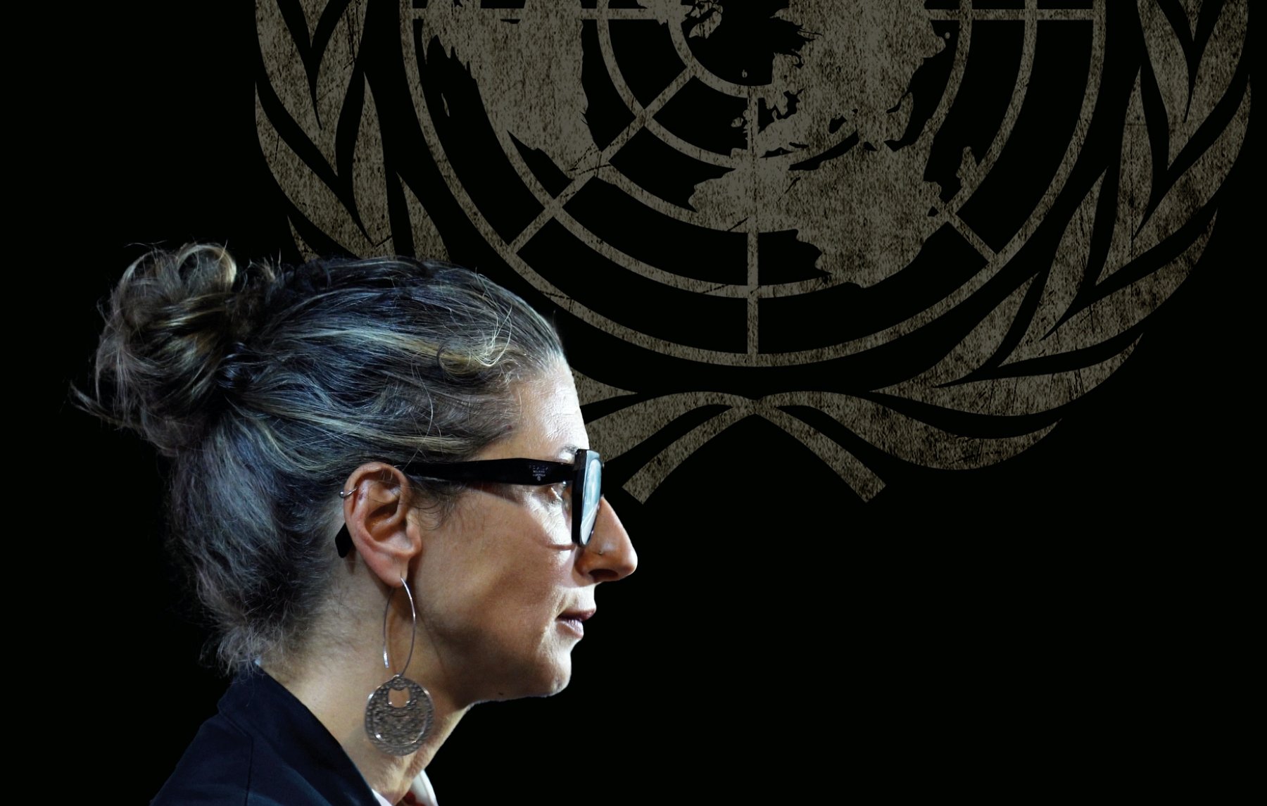 زنی با موهای خاکستری و عینک، در کنار نماد سازمان ملل، در حال صحبت کردن است.