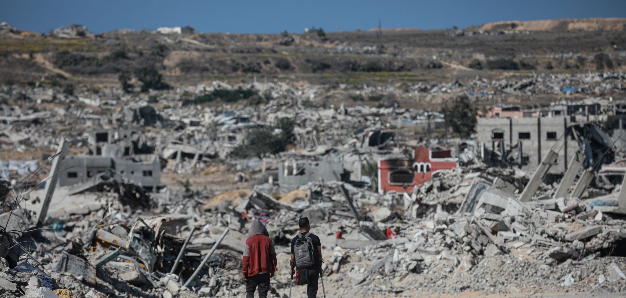 Two people walk through a vast landscape of destruction and rubble, with collapsed buildings around them.