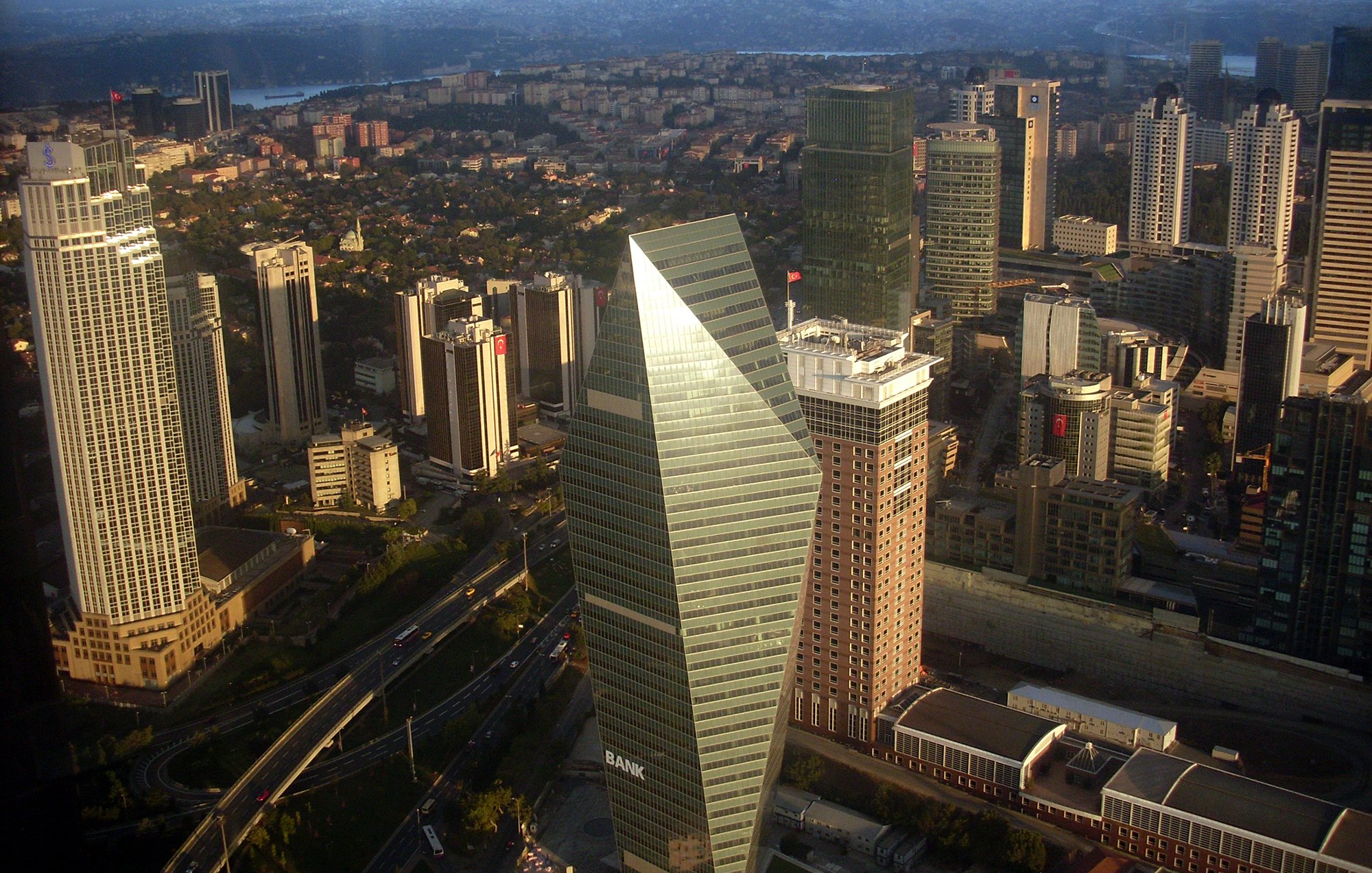 The image depicts a cityscape with a collection of modern skyscrapers and buildings. In the foreground, a distinctive triangular-shaped building stands out, likely serving as a bank or a financial institution. Surrounding it are various other high-rise buildings, indicating a bustling urban environment. The scene appears to be taken from a high vantage point, as it provides a wide view of the city, including green areas and roads. The skyline is clear, suggesting a bright day, with distant mountains or hills visible in the background.