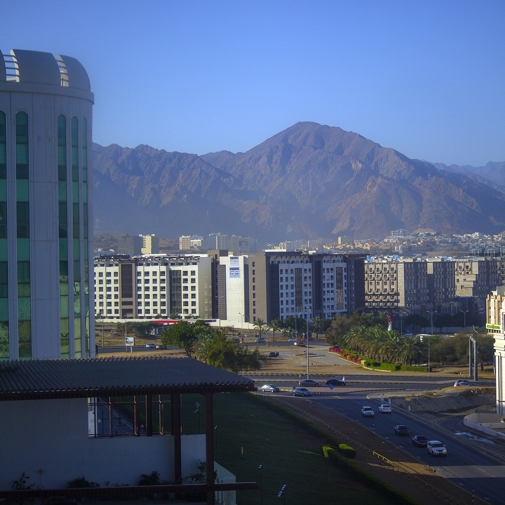 La imagen muestra un paisaje urbano con una mezcla de edificios modernos y montañas al fondo. A la izquierda, se puede ver un edificio de gran altura con un diseño contemporáneo. En el centro, hay una serie de edificios de varios pisos, que parecen ser parte de un área urbana. Al fondo, se aprecian montañas con una forma escarpada y un color terroso, indicando un ambiente montañoso. La luz del sol ilumina la escena, generando un ambiente claro y fresco. En primer plano, hay una carretera que conecta los diferentes sectores de la ciudad.